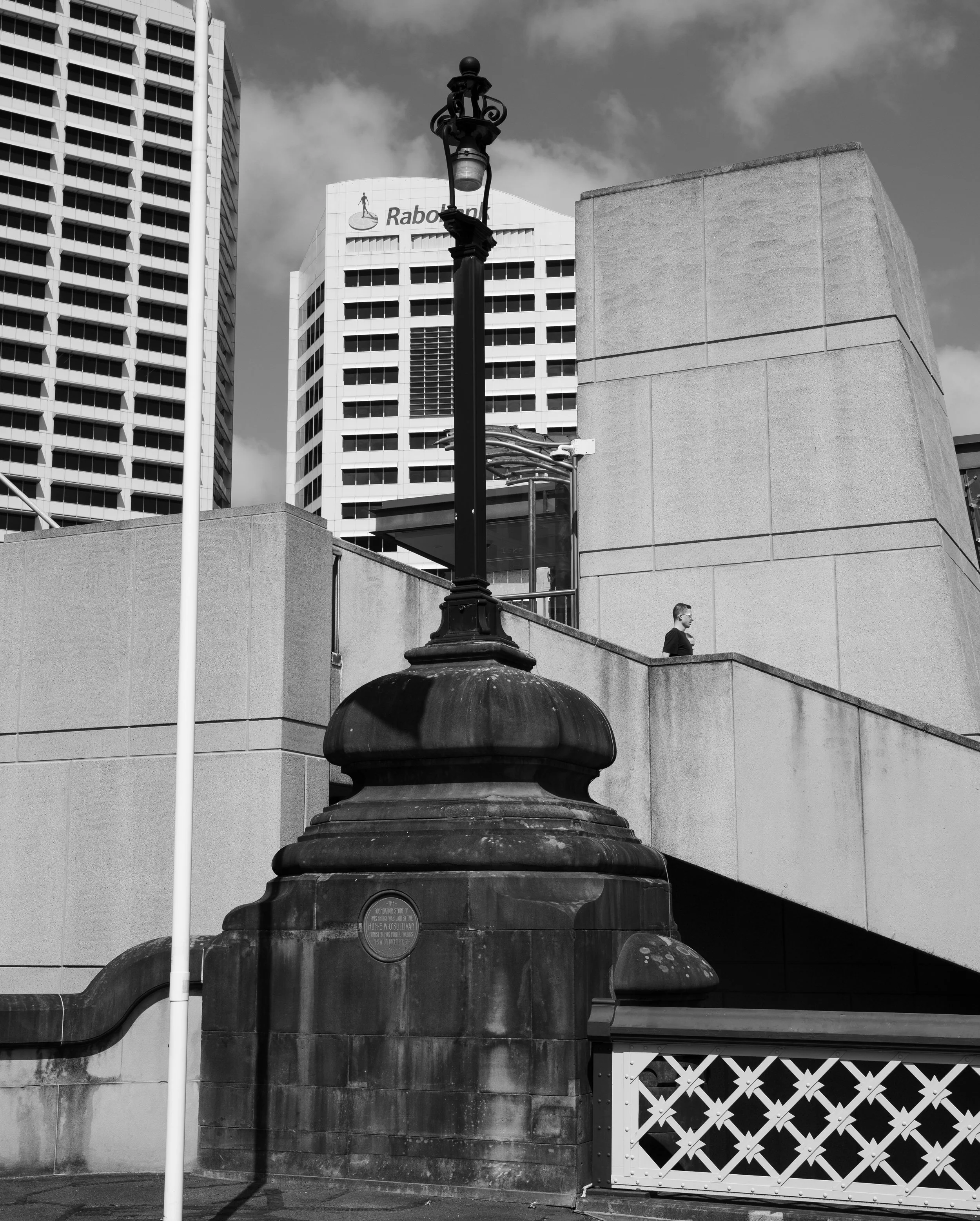 Black and white photo of a cityscape showing tall buildings, a decorative street lamp, the Rabobank building in the background, and a person walking on an elevated walkway.