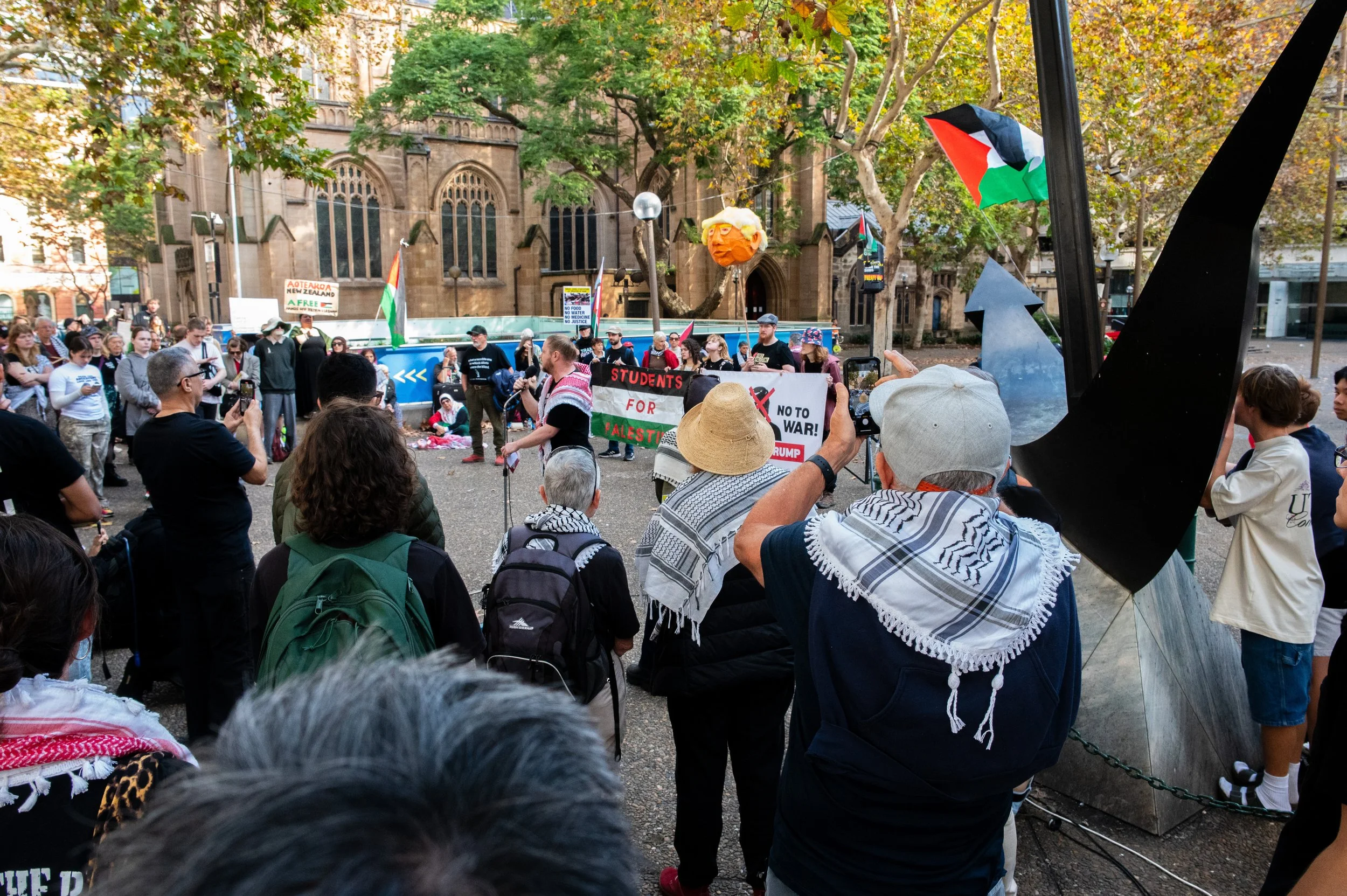 A crowd gathered at a protest or rally in front of a historic stone building with gothic windows, featuring people holding signs and Palestine flags, with some taking photos or videos, amid trees with autumn leaves.