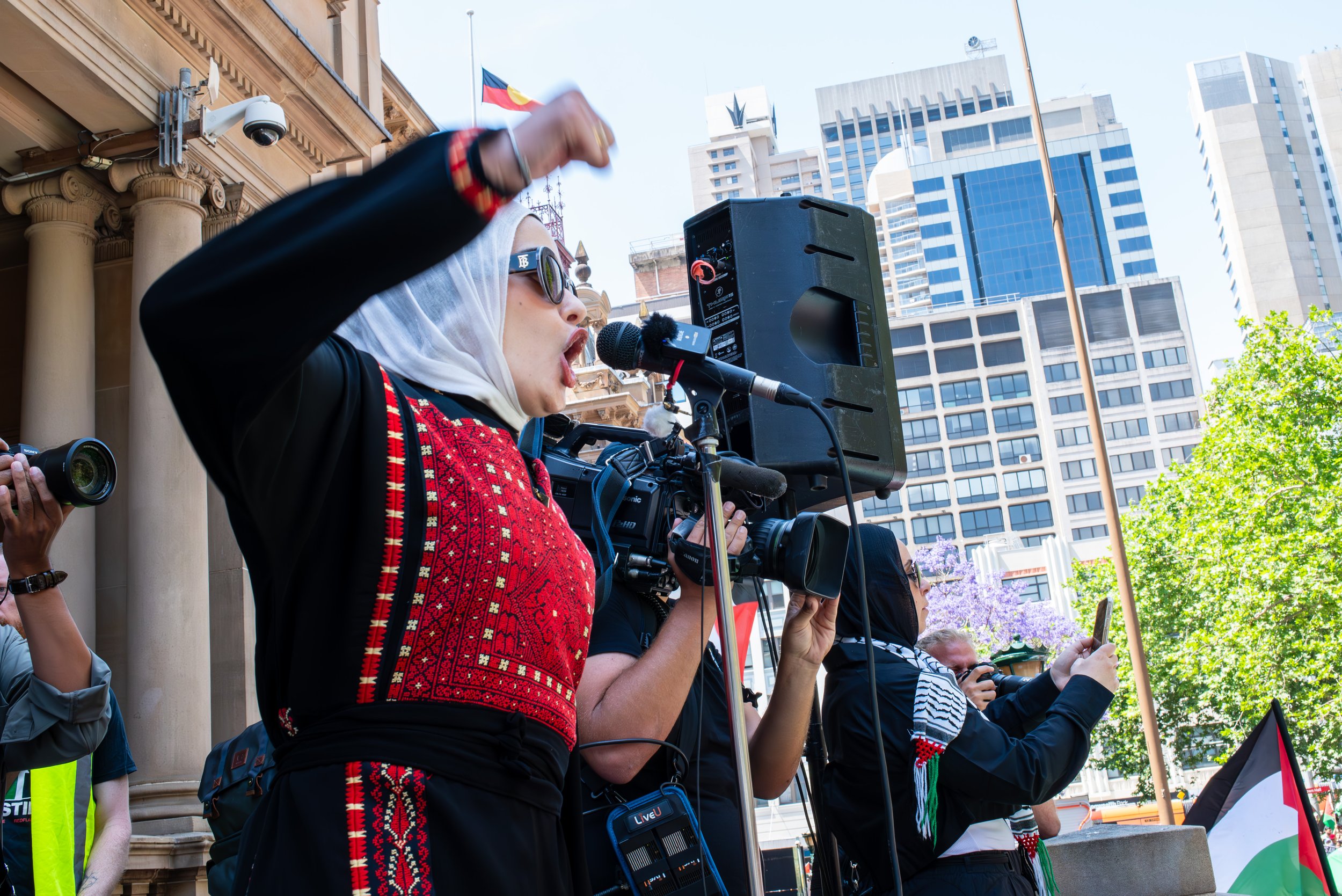 Woman in hijab and sunglasses speaking into microphone during outdoor protest, with cameras and a large speaker, city buildings and a flag in the background.