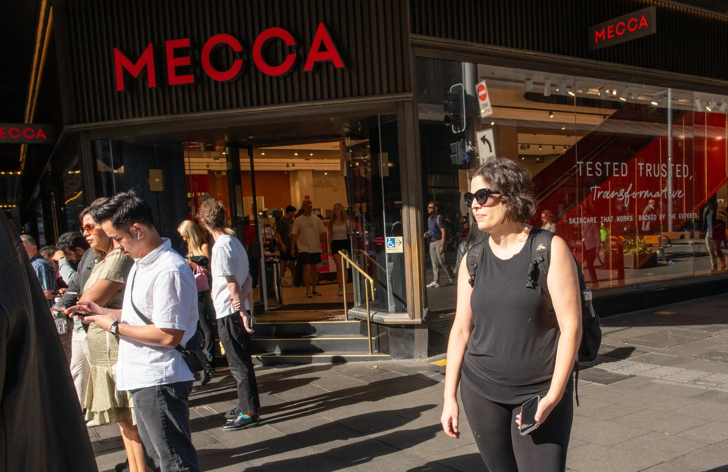 People standing outside a store named MECCA on a sunny day, some using their phones and wearing sunglasses.