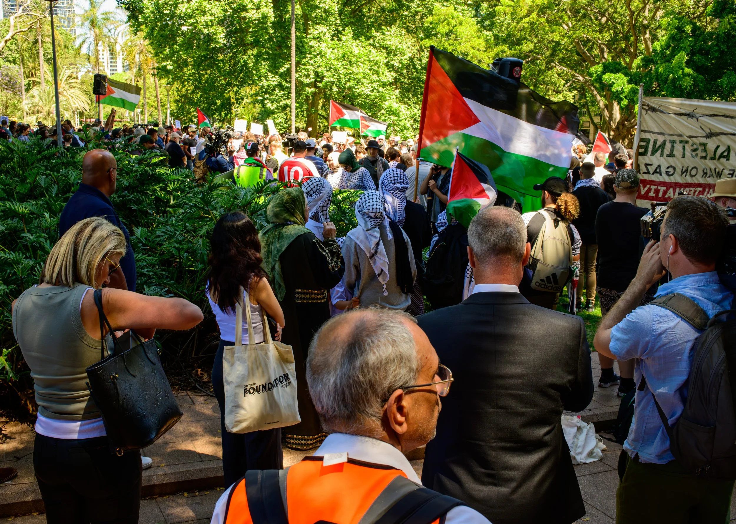 Crowd of people gathered in a park during a protest, holding Palestinian flags and signs with greenery and trees in the background.