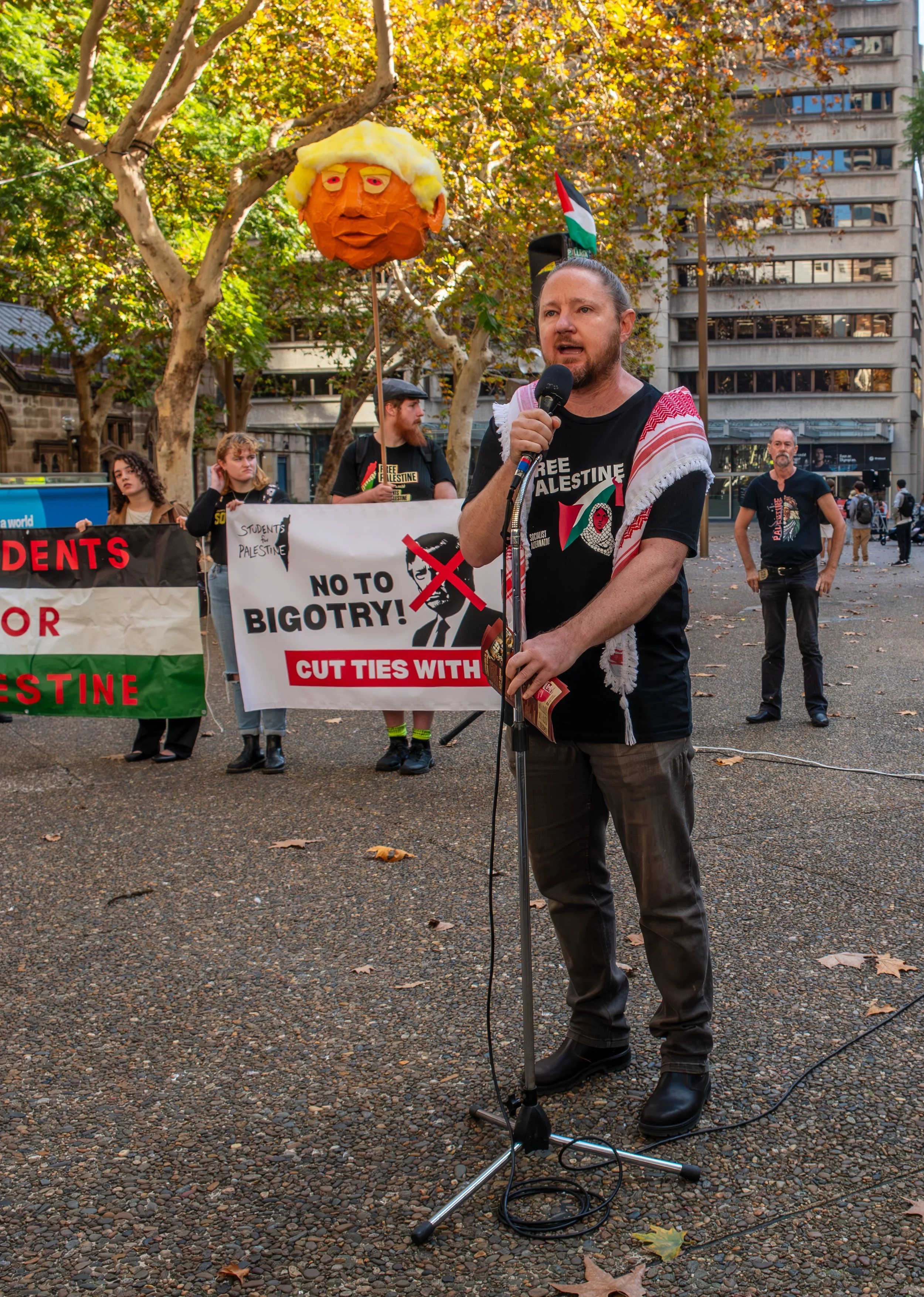 A man speaking into a microphone at a protest rally with Palestinians in the background holding banners that say 'No to Bigotry! Cut ties with' and 'Students for Palestine'. One banner also features a caricature of a person with a red cross over the face. A balloon resembling Donald Trump with his distinctive hair is hanging overhead. The protest is taking place in an outdoor urban setting with trees and buildings visible.