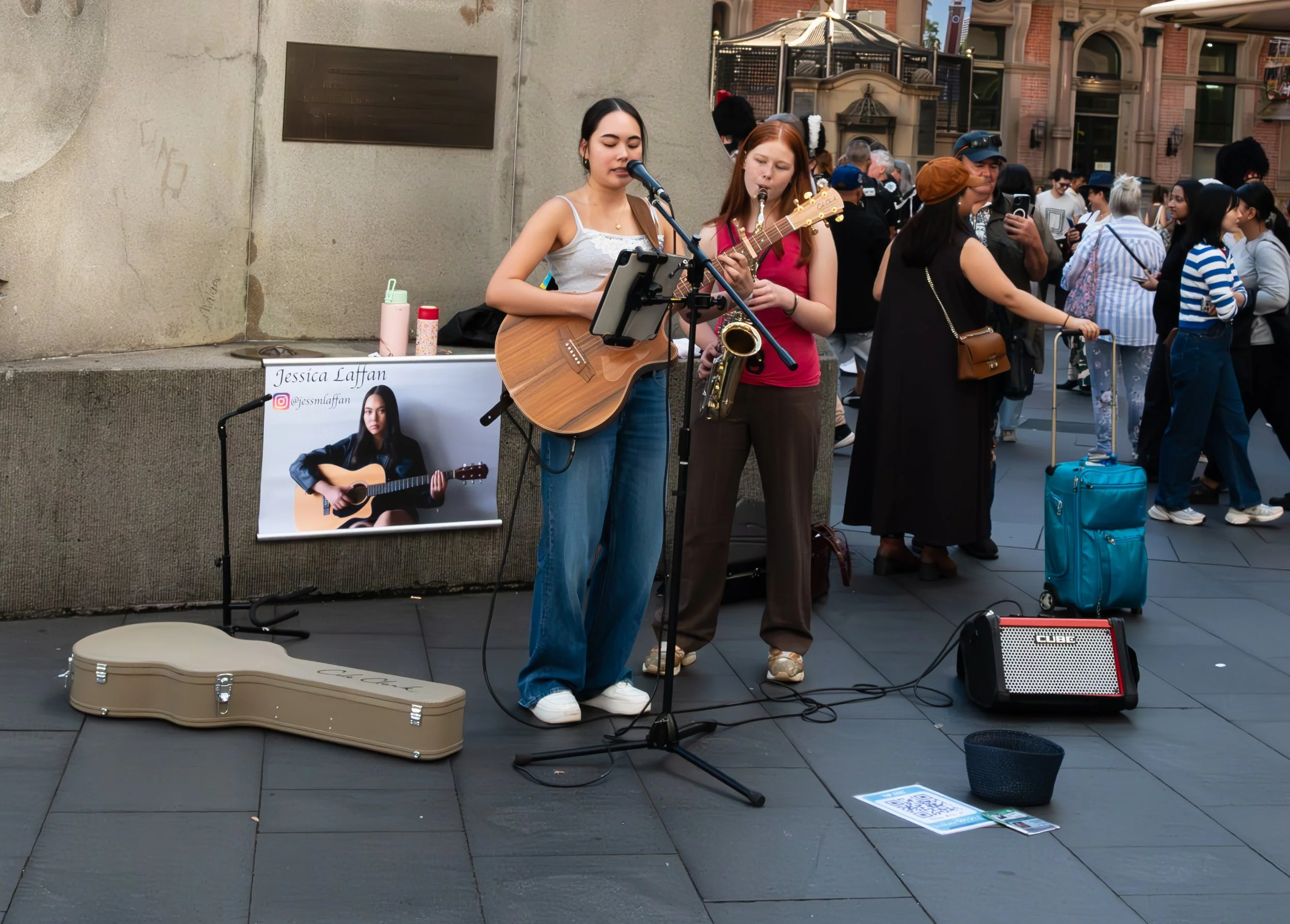 Two young women performing street music with one playing guitar and singing into a microphone, the other playing a saxophone, in a crowded outdoor urban area. There is a poster of Jessica Laffan on the wall beside them, a guitar case on the ground, and a small amplifier nearby, with a diverse crowd of people in the background.