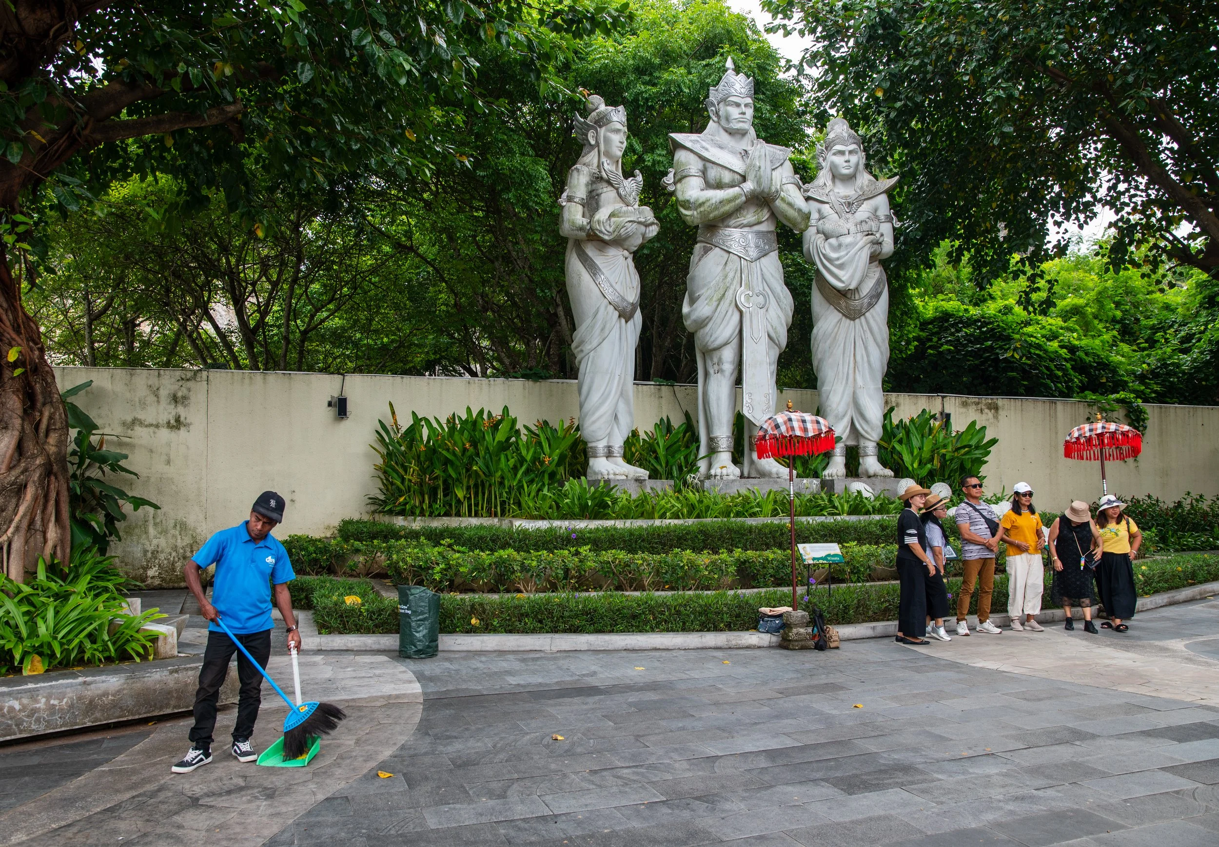 Group of tourists standing near a white wall with a large granite monument of three mythological figures, surrounded by green trees and shrubs.
