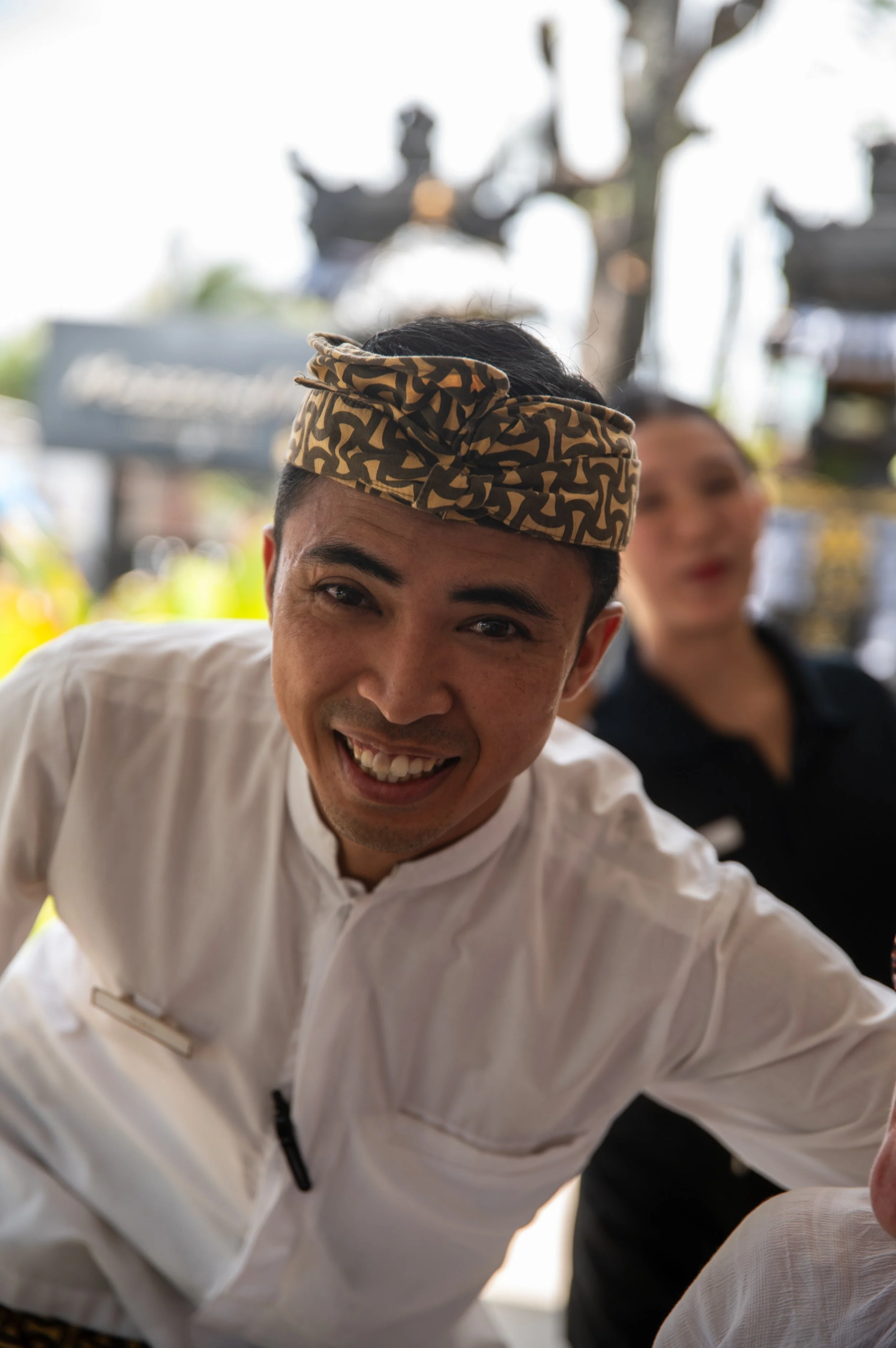Close-up of a smiling man wearing a patterned headband and white chef's coat, with a woman blurred in the background.