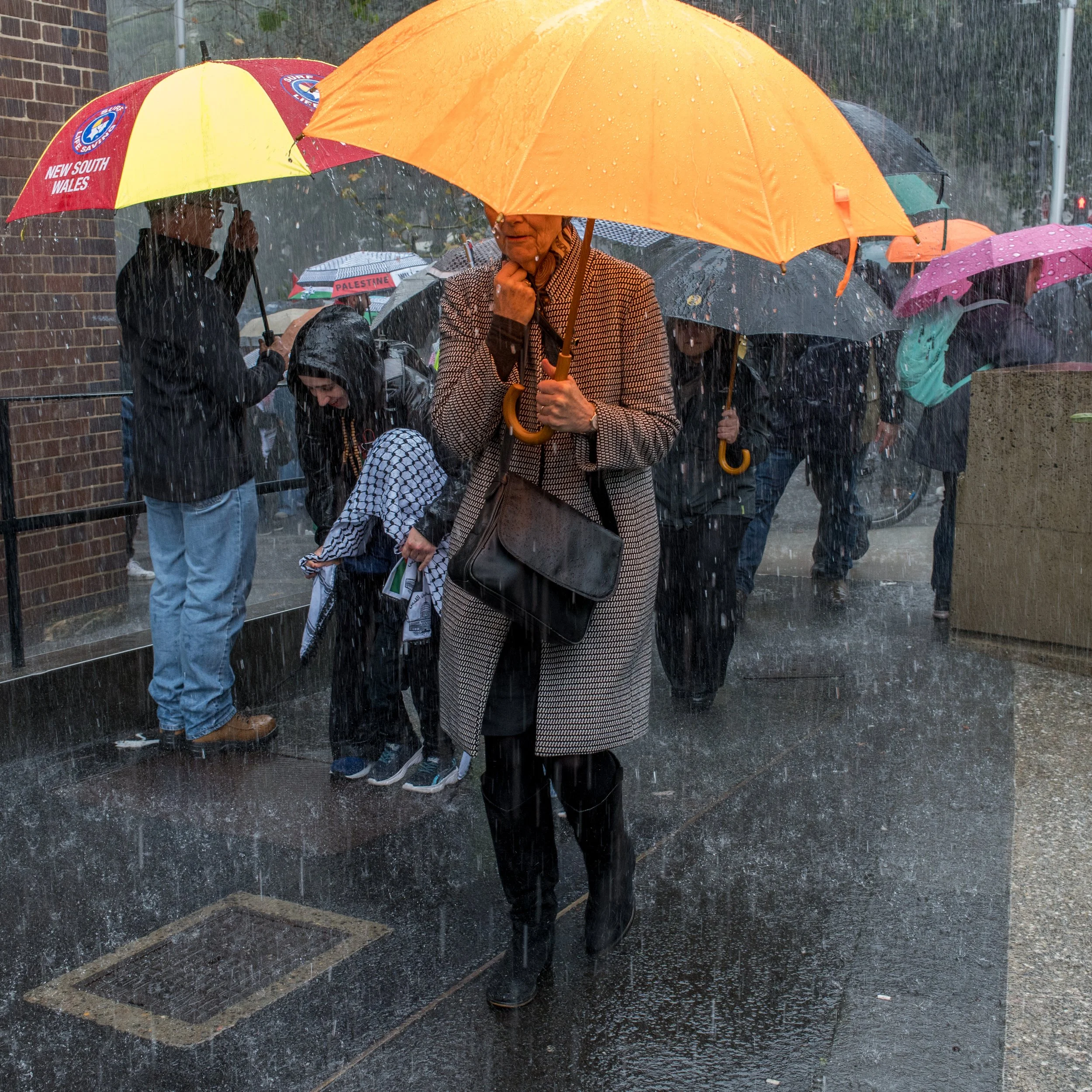 People standing in the rain with umbrellas on a city sidewalk, including a woman in a coat and boots, appearing wet from the rain.