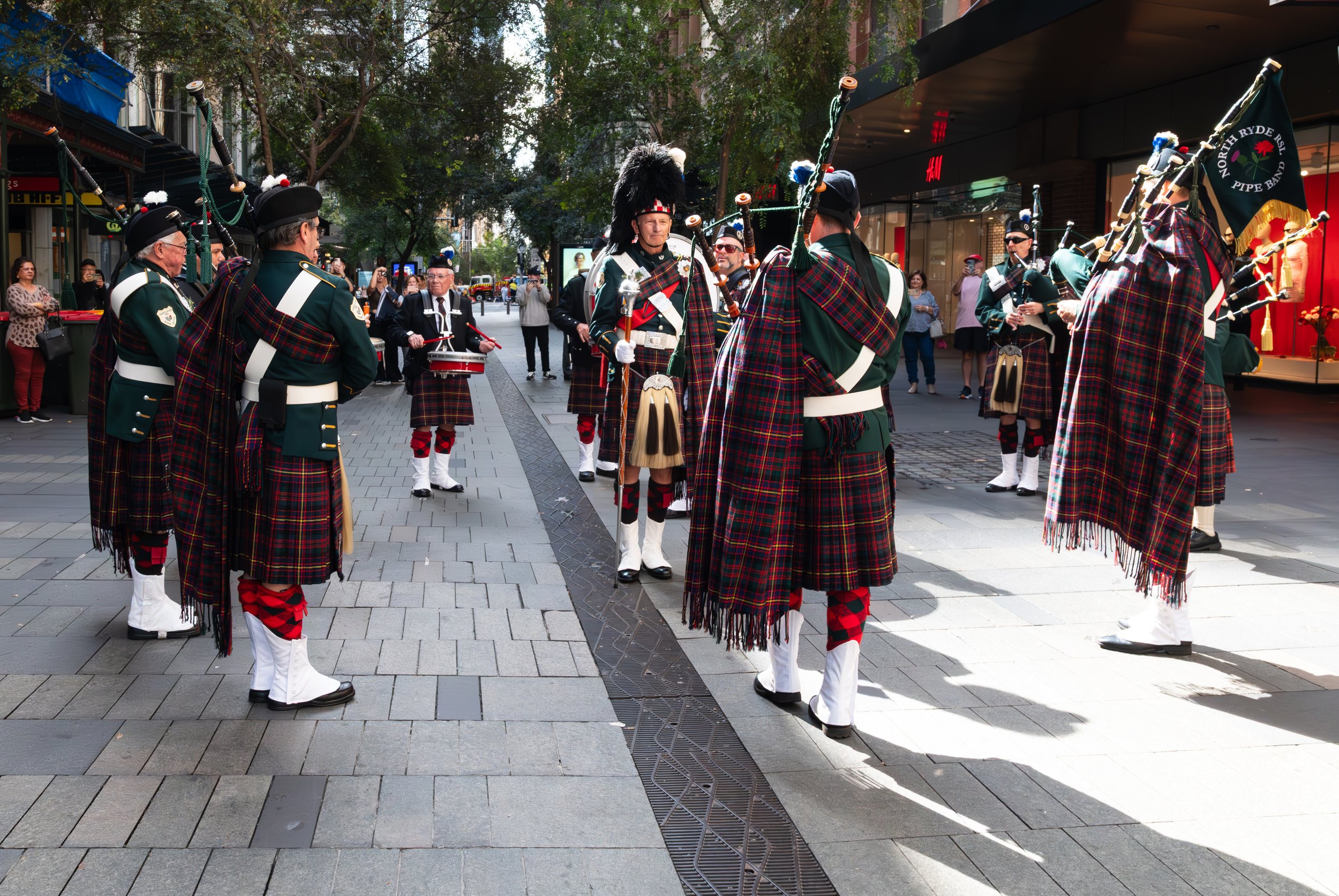 A traditional Scottish pipe band dressed in green jackets, tartan kilts, and black hats performing on a city street, with spectators watching.