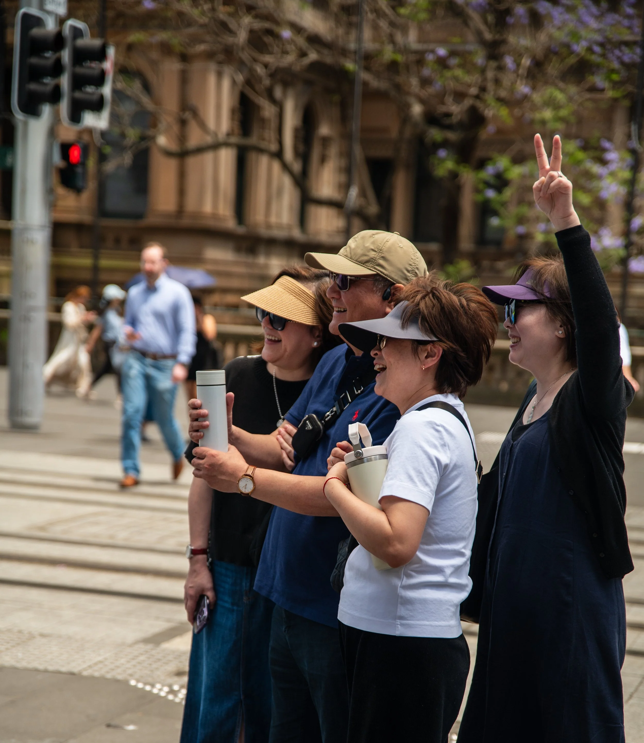 Group of five cheerful people, four women and one man wearing hats and sunglasses, posing for a photo on a city street, with a woman on the far right making a peace sign with her fingers. In the background, there are traffic lights and a historic-looking building with purple flowering trees.