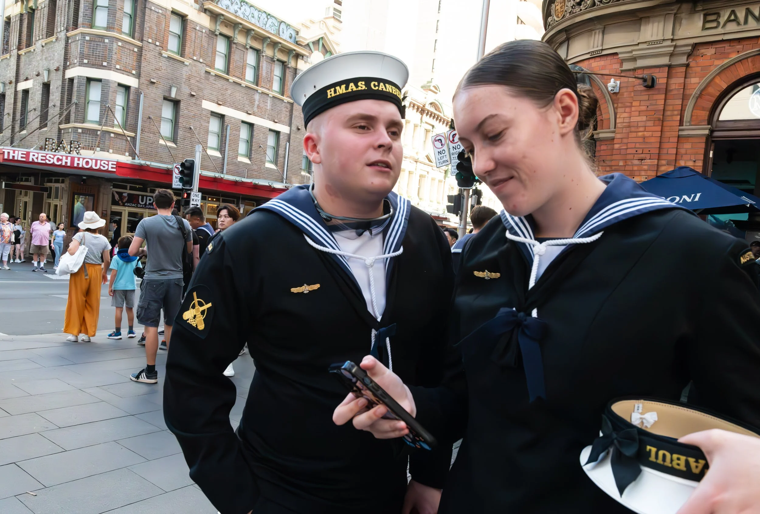 Two young people in Navy uniforms standing on a city street looking at a phone, with several pedestrians in the background.
