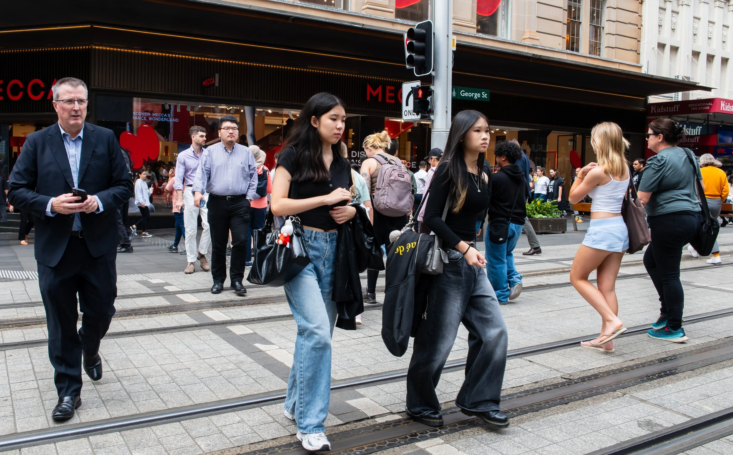 People crossing a busy city street at a crosswalk, with storefronts and a traffic light in the background.