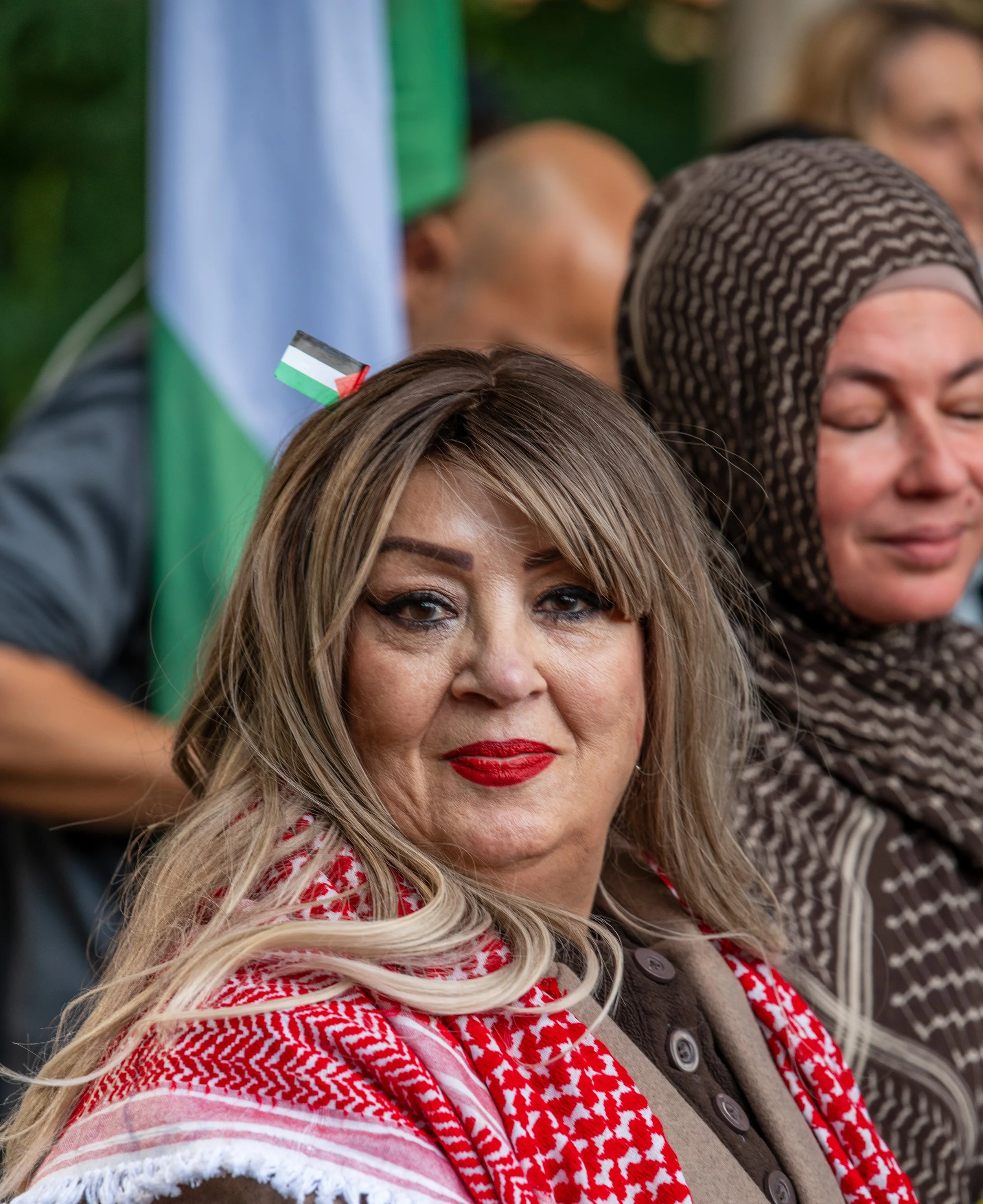 A woman with blonde hair, red lipstick, and eye makeup, wearing a red and white patterned scarf, sitting with others in a crowd at an outdoor event, with a flag in the background.