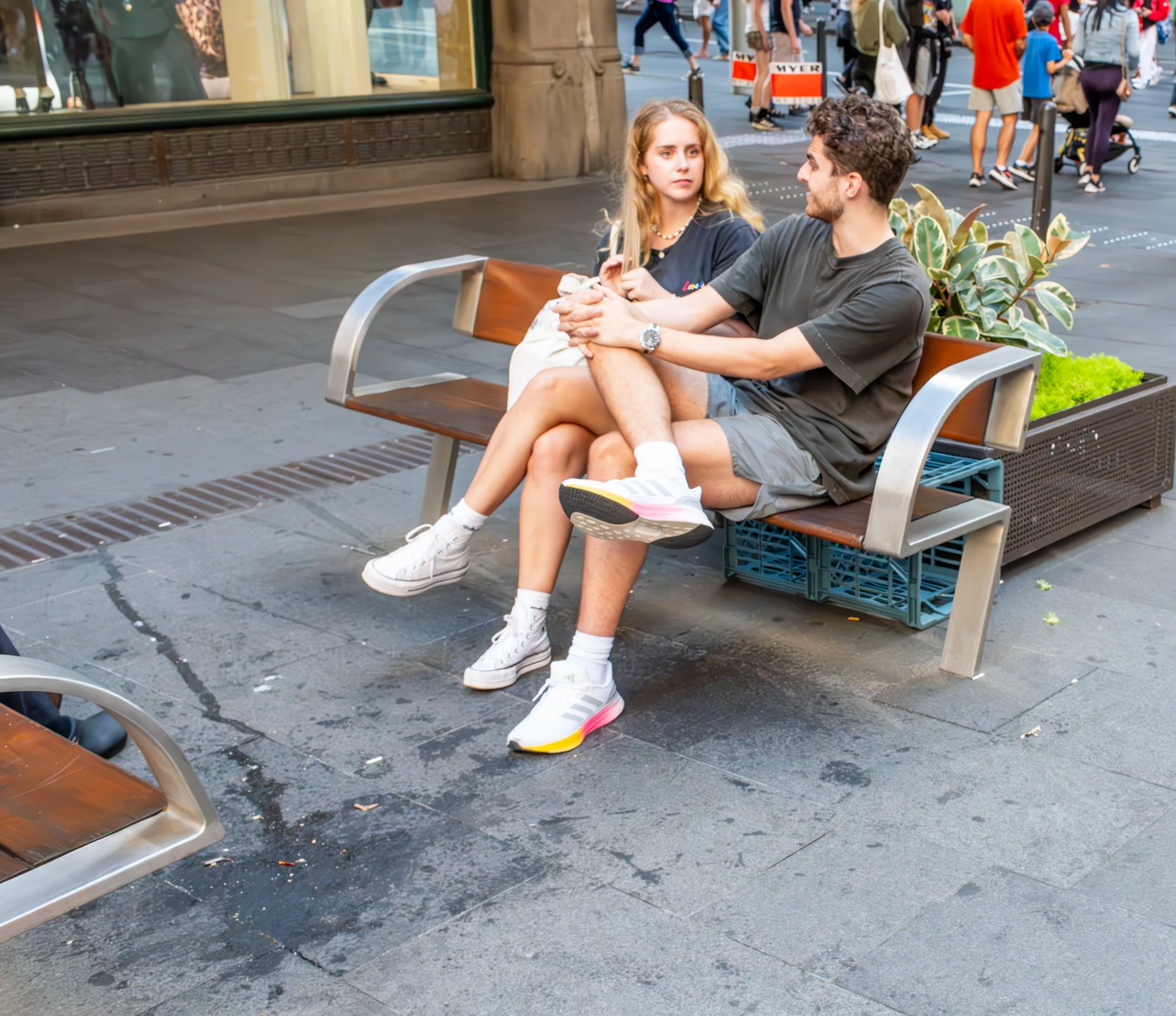 A young man and woman sit on a city park bench, with the man holding the woman's legs, engaging in conversation. They are wearing casual clothes, and the bench has green plants behind them. In the background, there are people walking on the busy street.