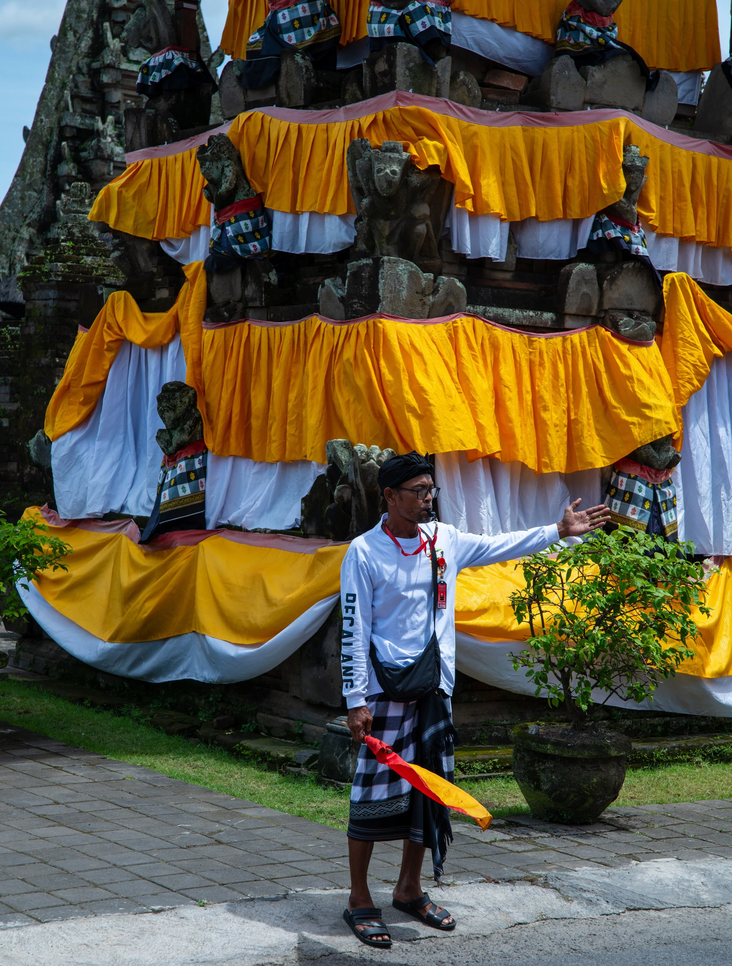 A person dressed in traditional clothing with a cloth wrapped around their waist, holding a red and yellow object, standing outdoors near a structure decorated with yellow and white fabric and stone sculptures.