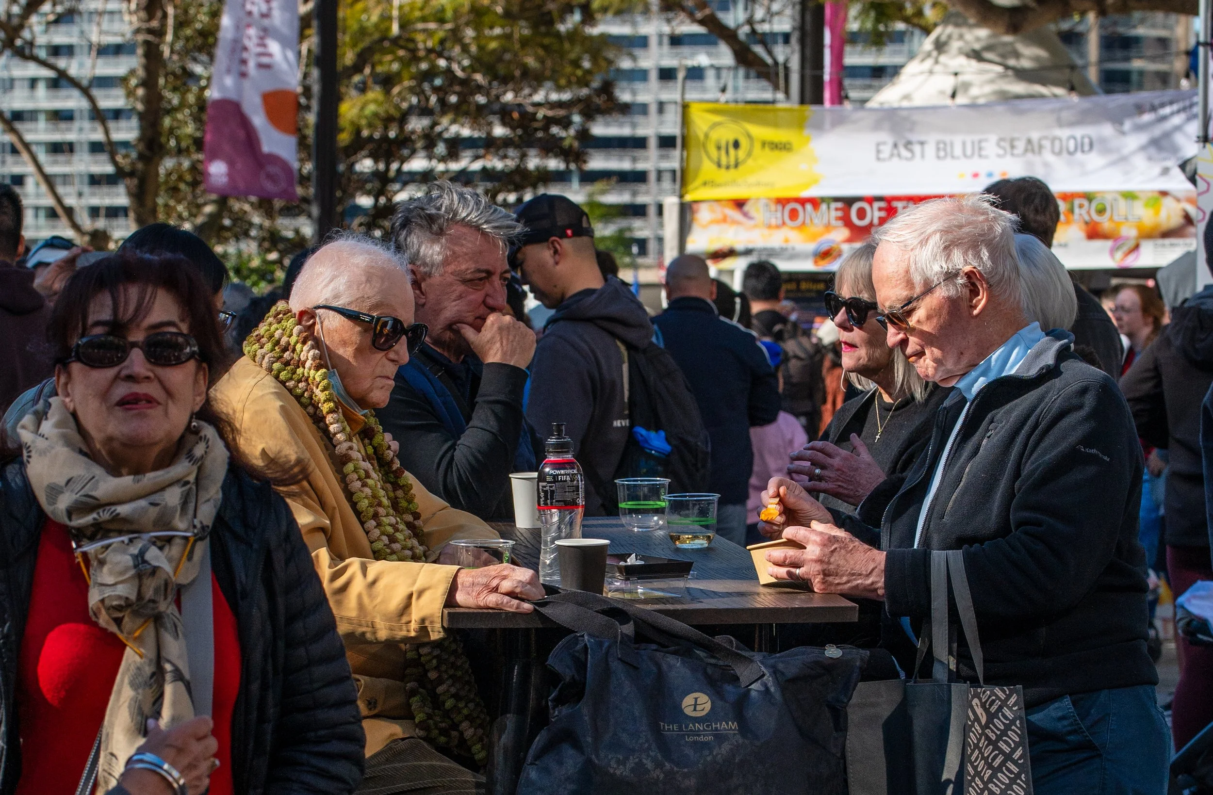 A group of elderly and middle-aged people gathered around a high table at an outdoor event, some wearing sunglasses, with food and drinks on the table, and tents and banners in the background.