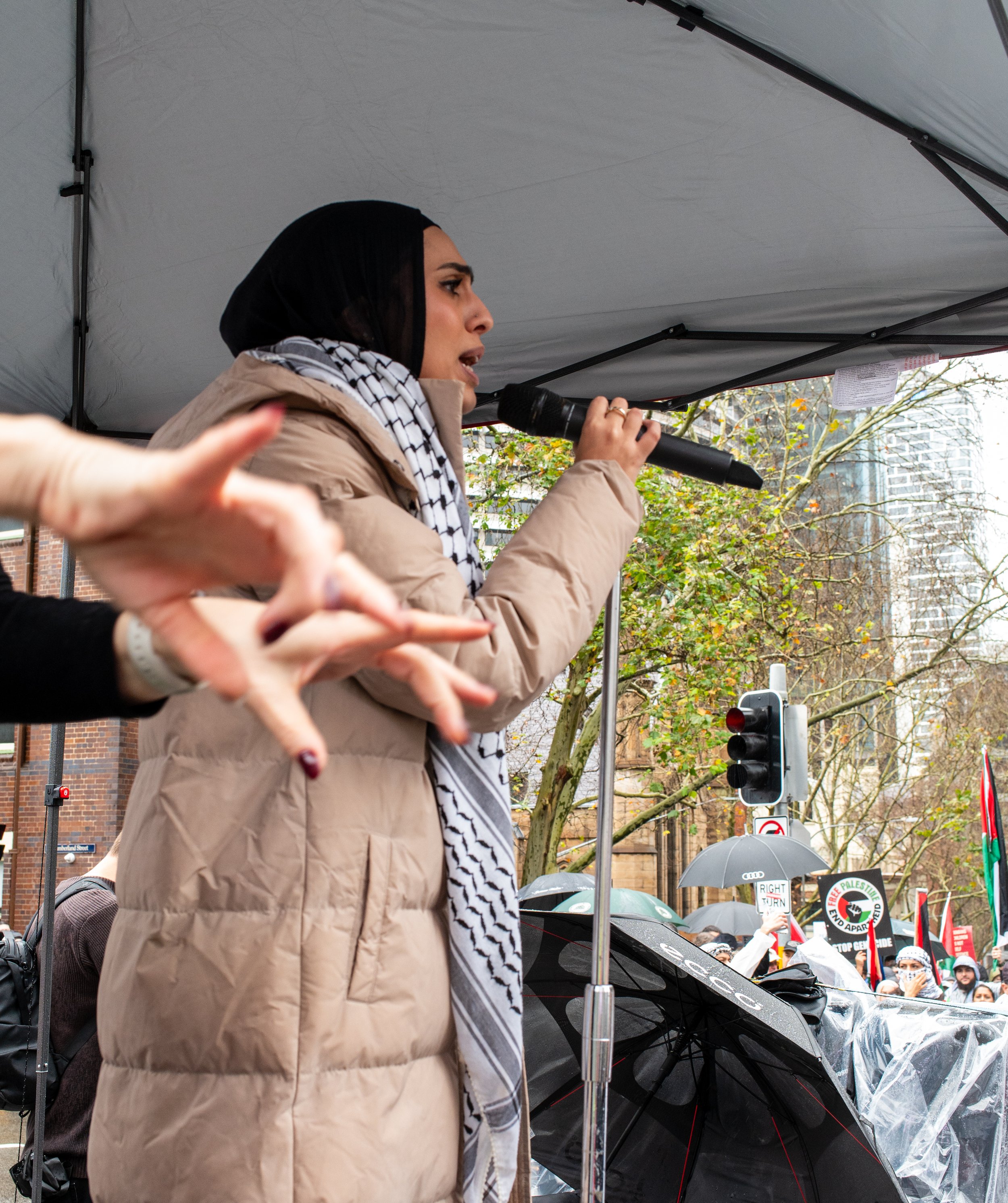 A woman wearing a beige jacket and a black and white keffiyeh scarf speaks into a microphone at a protest or rally, with a crowd holding signs and umbrellas behind her.