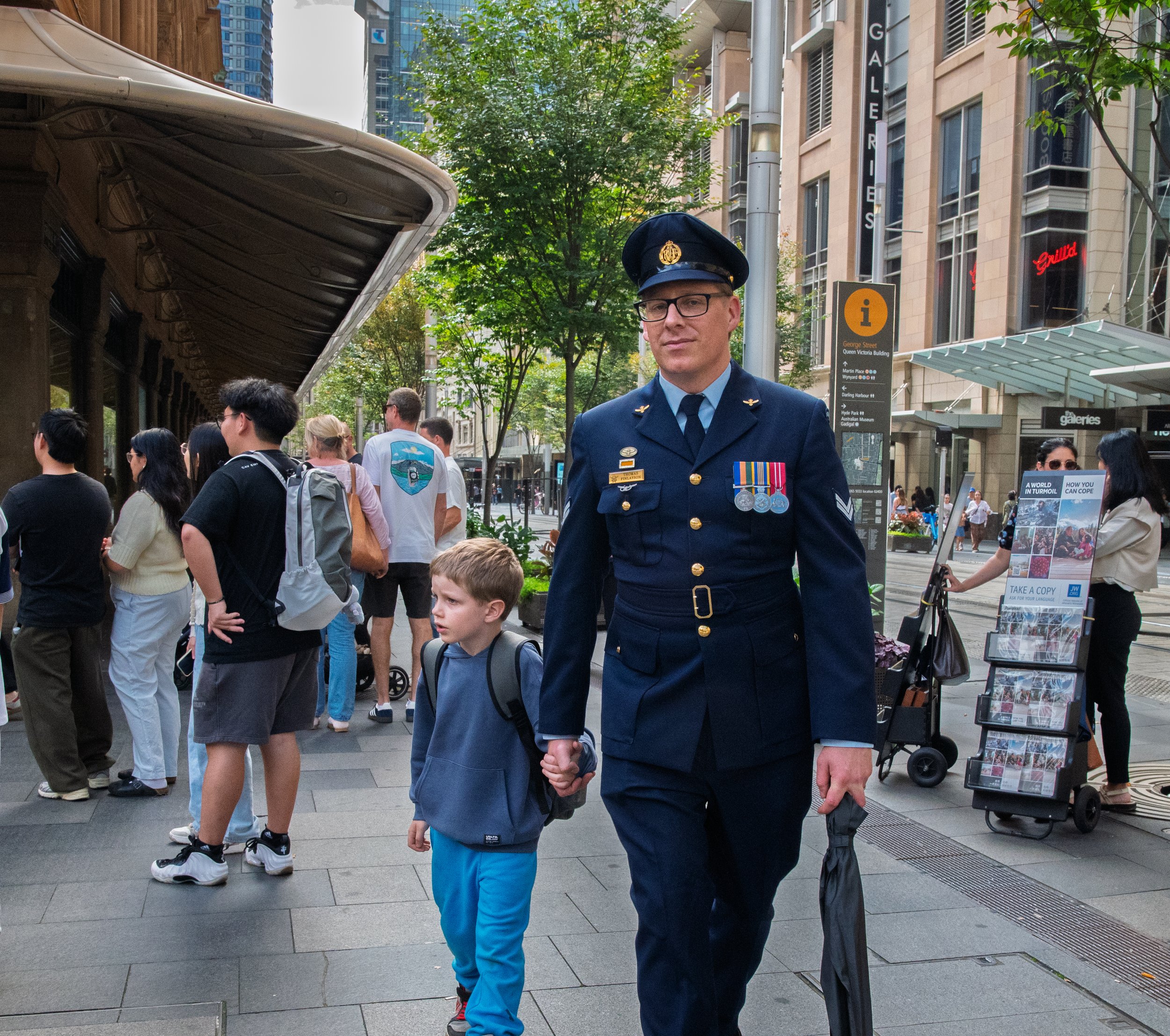 A police officer in uniform holding hands with a young boy on a busy city sidewalk, with people waiting in line and storefronts in the background.