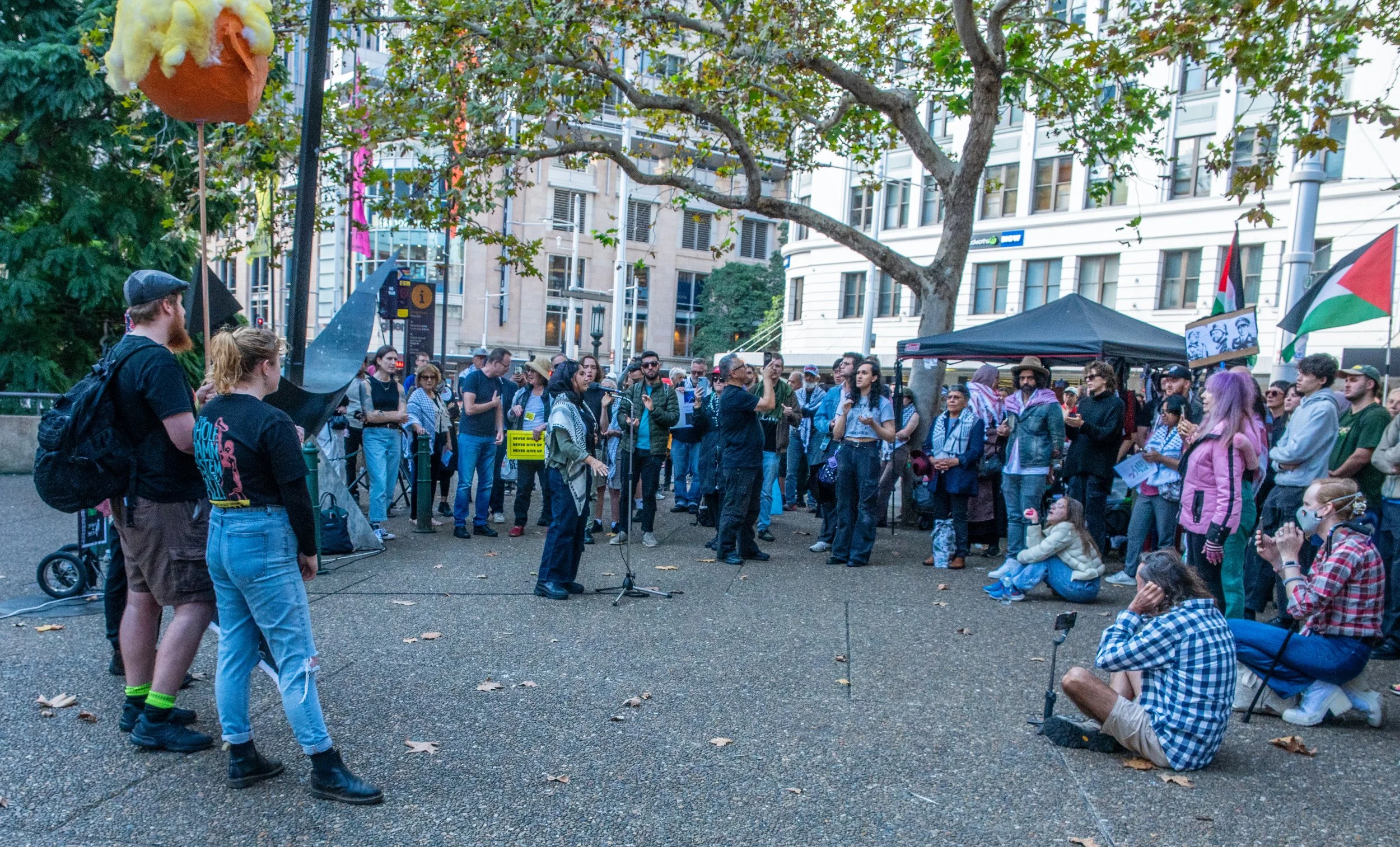 A group of people gathered outdoors for a protest or rally. They are standing and sitting, some holding flags and signs, with a speaker addressing the crowd. Trees and tall buildings are visible in the background.