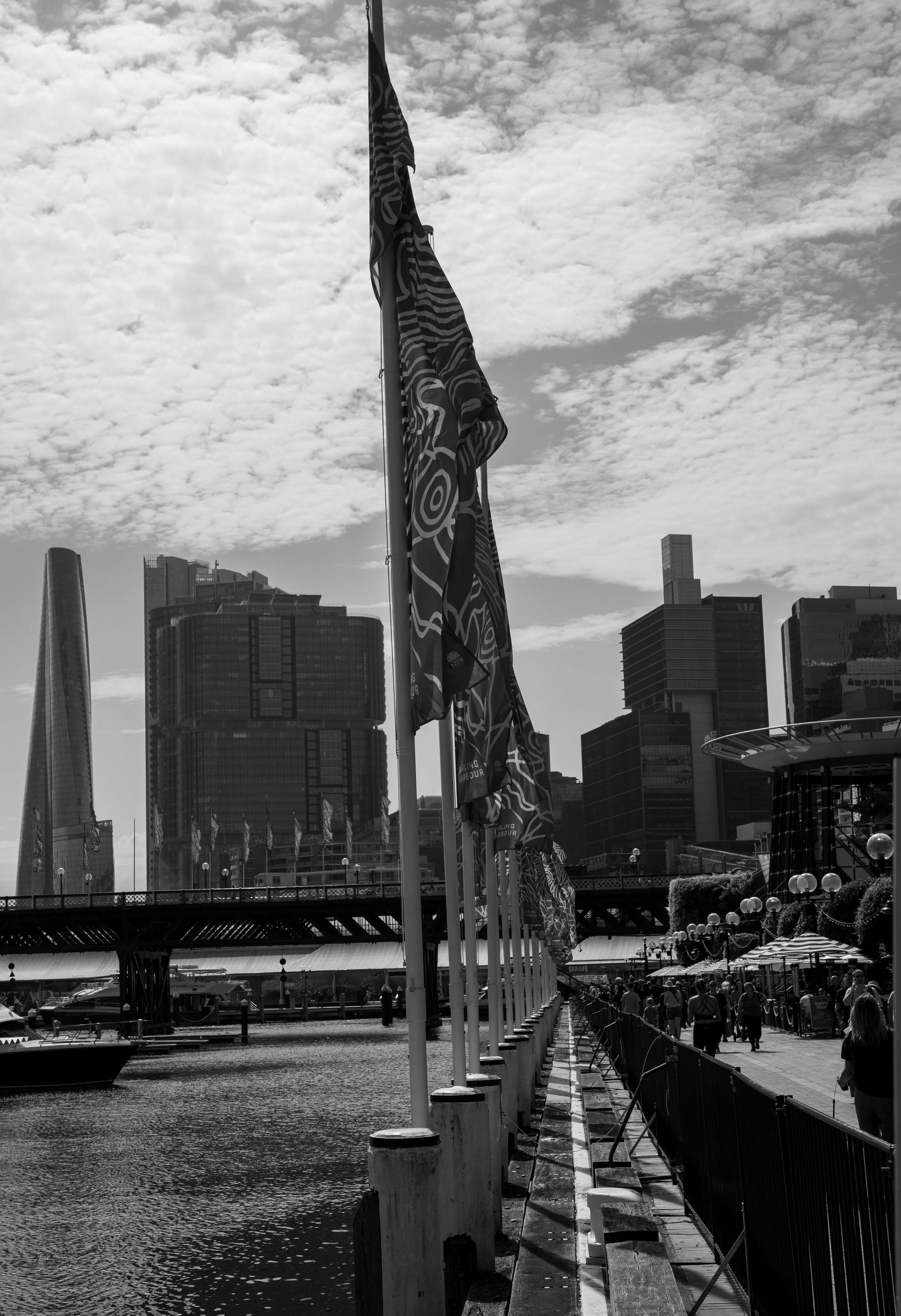 Black and white photo of a waterfront in a city with tall buildings, flagpoles with decorative flags on the dock, and people walking along the promenade.