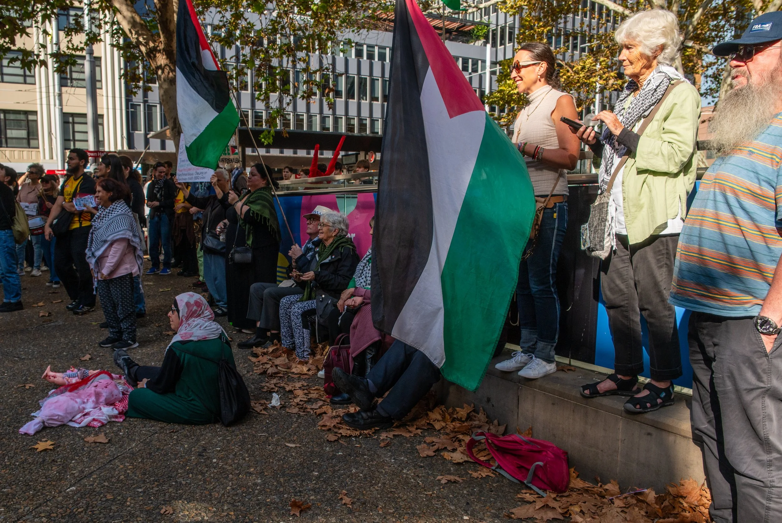 A diverse group of people gathered outdoors, some standing and some sitting, during a protest or rally. Several individuals hold Palestinian flags, and one person is sitting on the ground wearing a hijab and holding a small Palestinian flag. The background shows modern buildings and trees with autumn leaves on the ground.