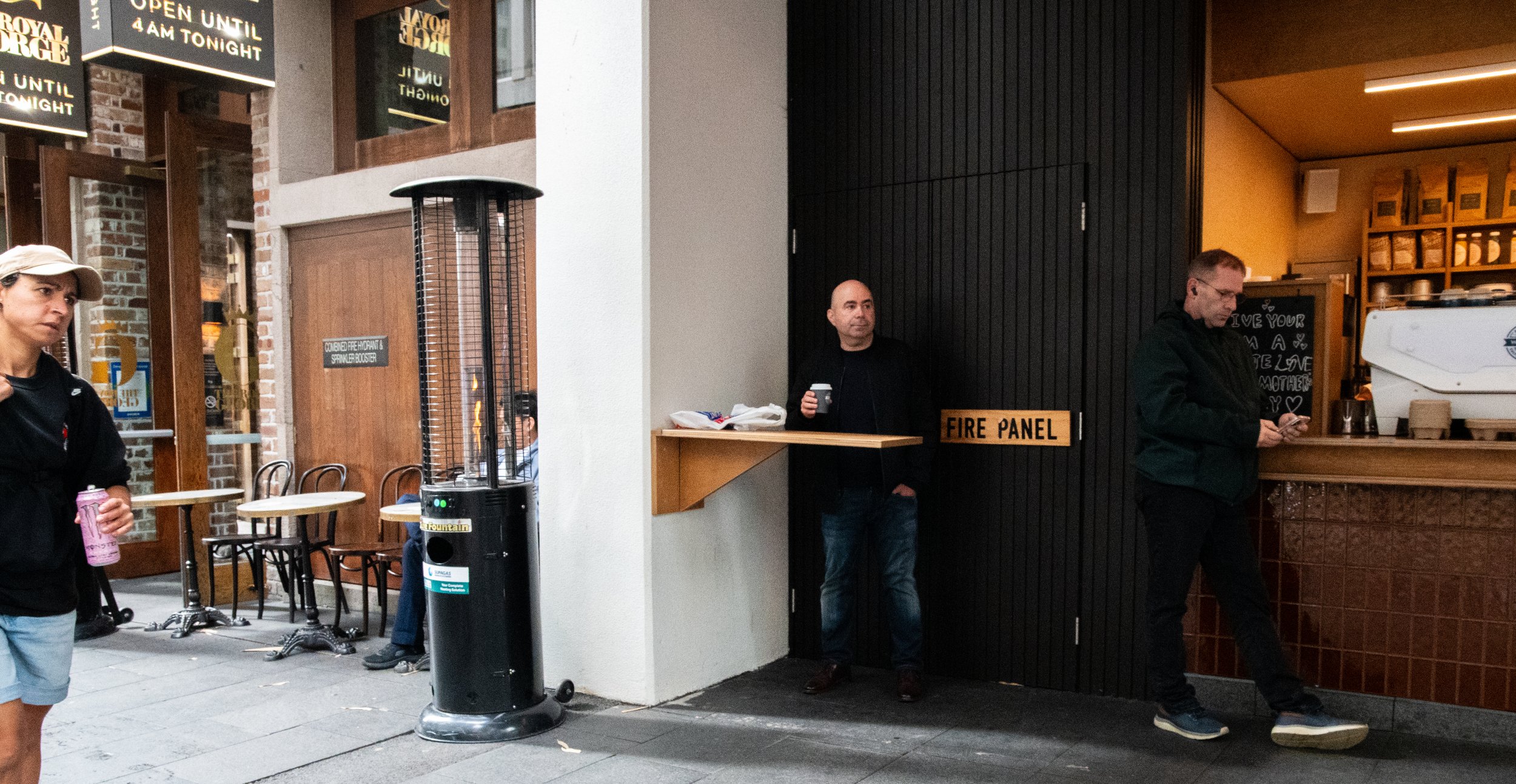 People standing near a coffee shop counter on a city sidewalk, with one person holding a coffee cup and another looking at their phone, outside tables and chairs are visible.