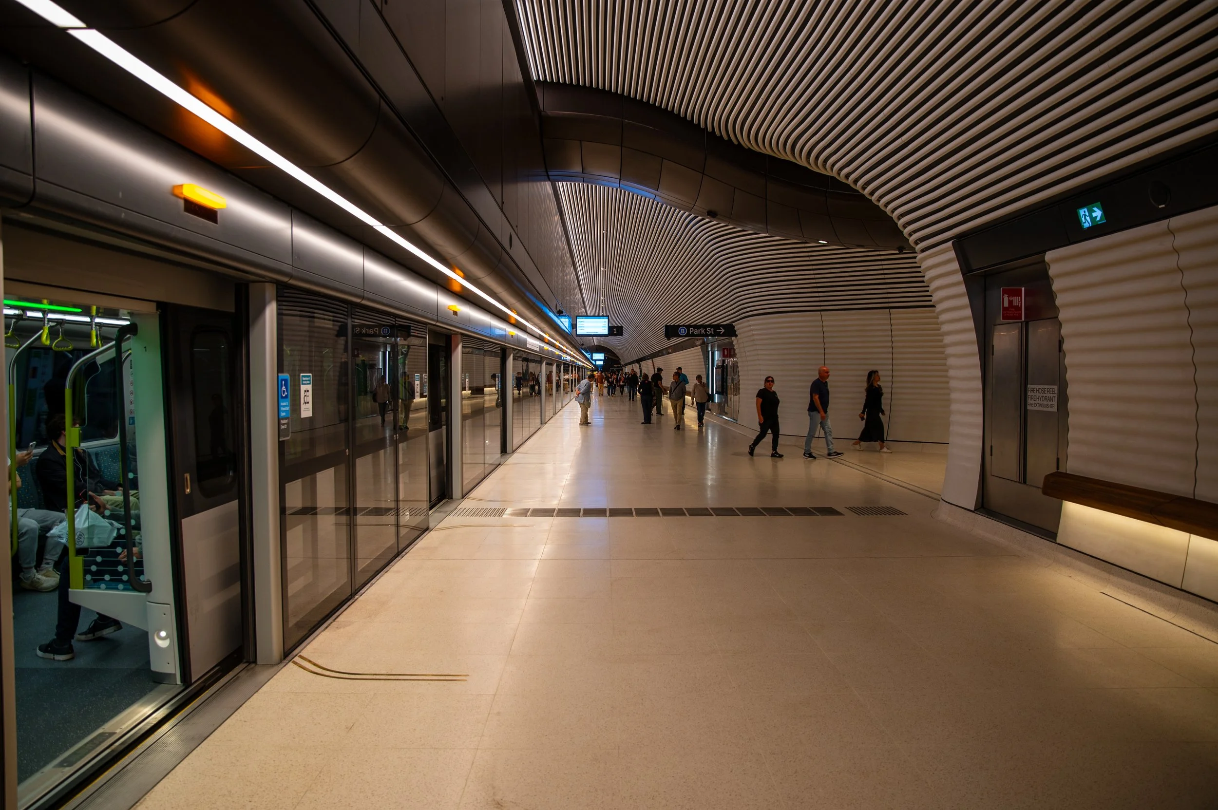 Empty underground train station with a curved modern ceiling, glass doors, and several people walking or standing.