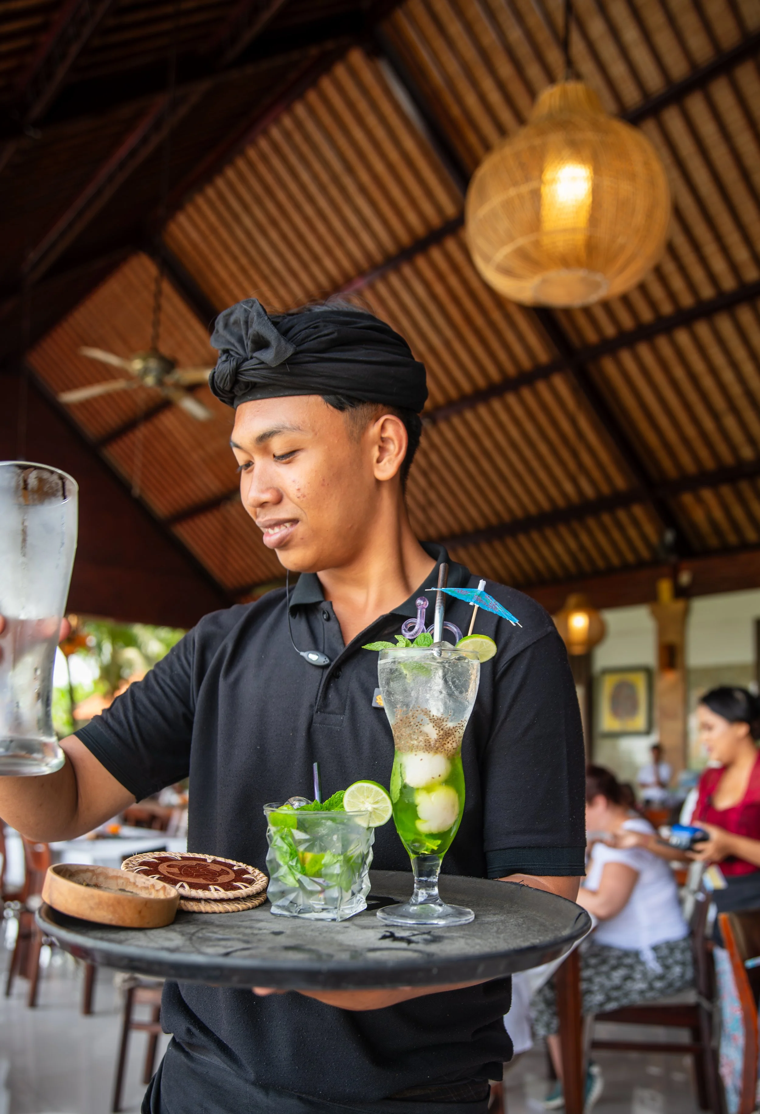 A young waiter with a black turban and uniform serving drinks at a restaurant. The drinks include a mojito with lime, mint, and ice, and a colorful cocktail with decorative umbrellas. The restaurant has a wooden ceiling with a hanging lamp and ceiling fan.