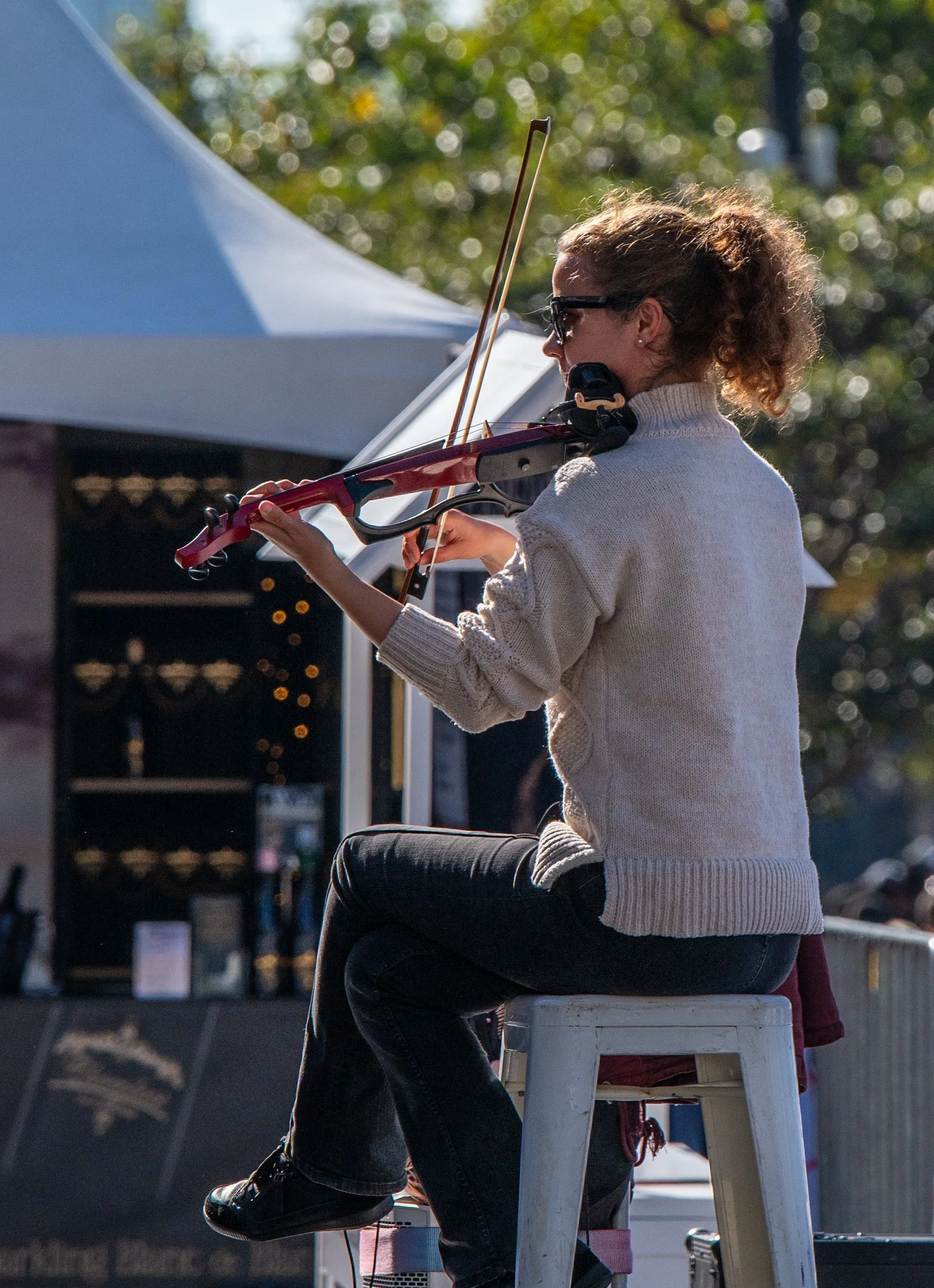 A woman playing the violin while sitting on a stool outdoors, wearing sunglasses and a cream sweater.