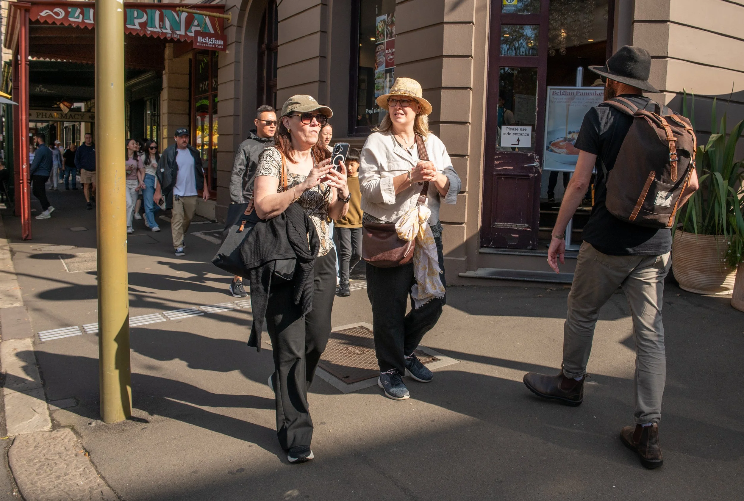 Three women and one man on a city sidewalk, with some women taking photos or talking and a man walking away, near shops and pedestrians in the background.