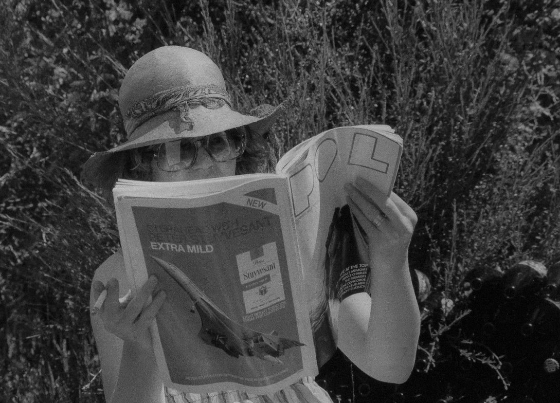Person wearing large hat and glasses reading a newspaper outdoors.