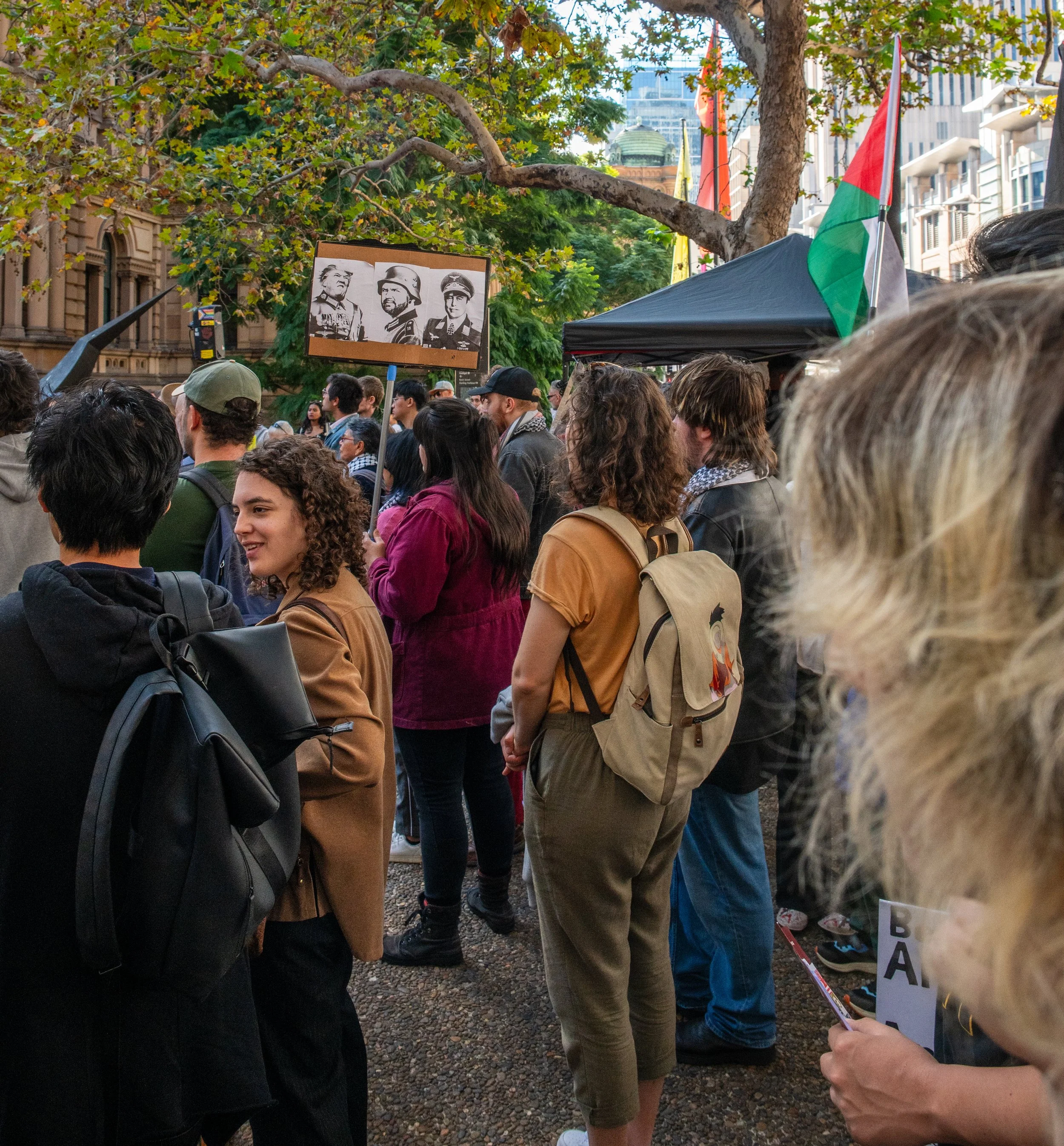 Crowd of people gathered outdoors under trees at a protest or rally, with a sign featuring historical military figures and a flag with red, green, black, and white colors in the background.