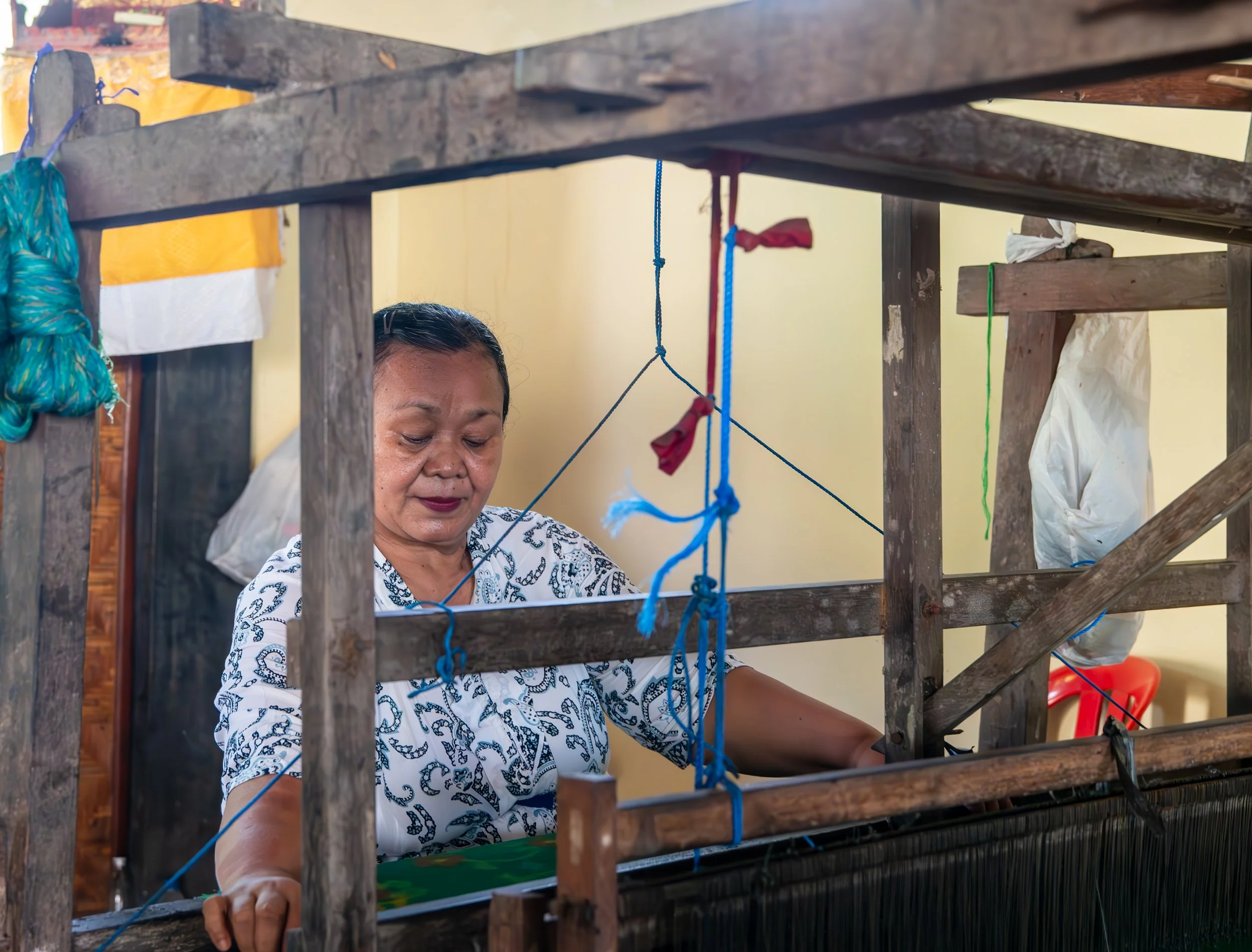 A woman with dark hair and wearing a white patterned shirt working on a craft project behind a wooden frame with colored strings tied on it.