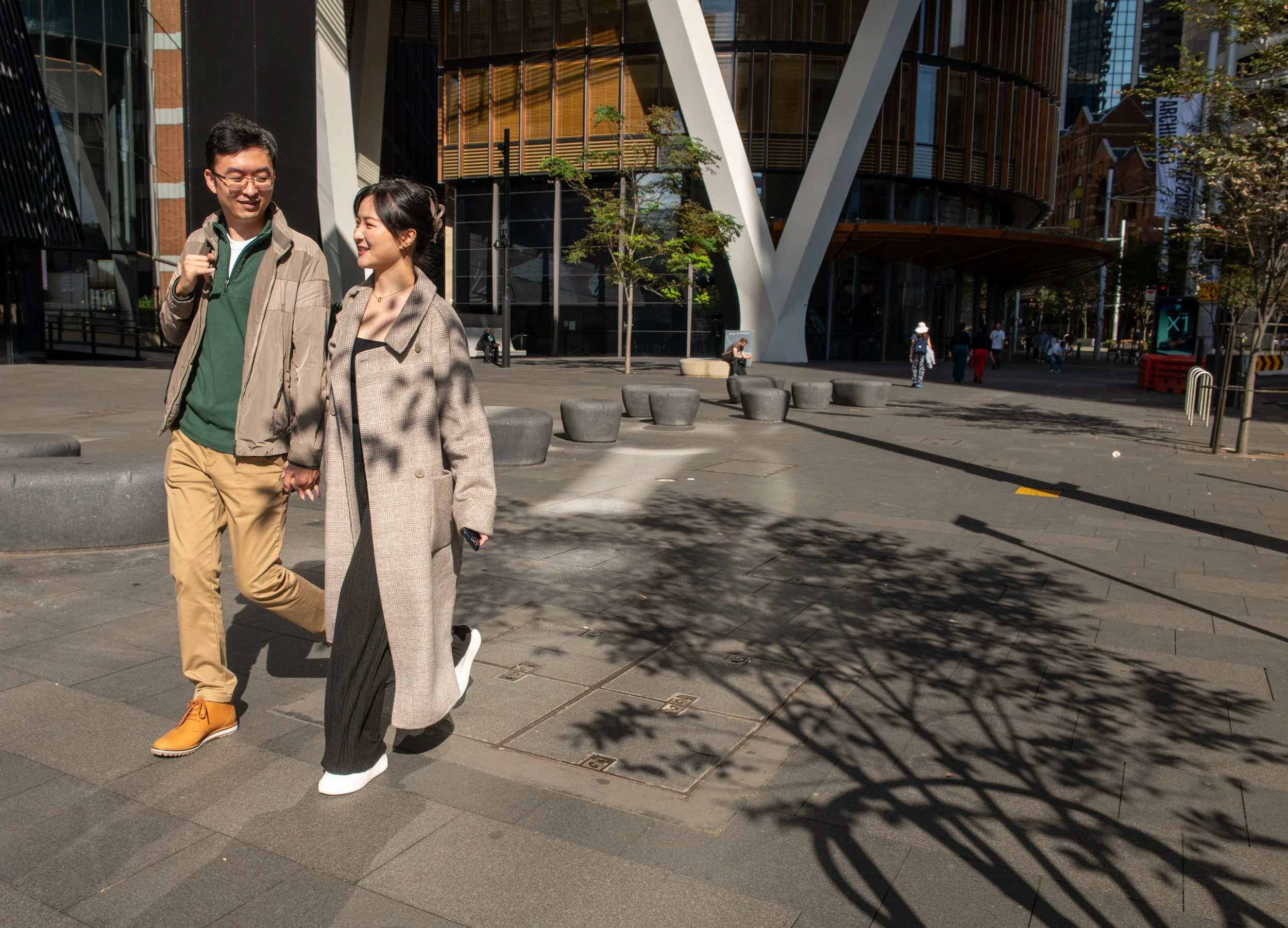 A young man and woman walking hand-in-hand on a city sidewalk on a sunny day, with modern buildings and a few trees in the background.