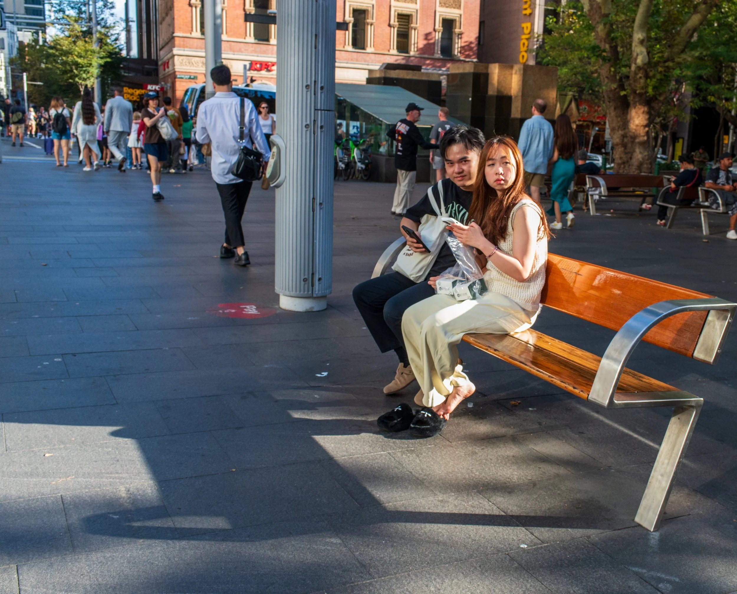 Two young adults sitting on a park bench in a busy city street, looking at their phones. There are many people walking in the background, and buildings with signs and trees lining the street.