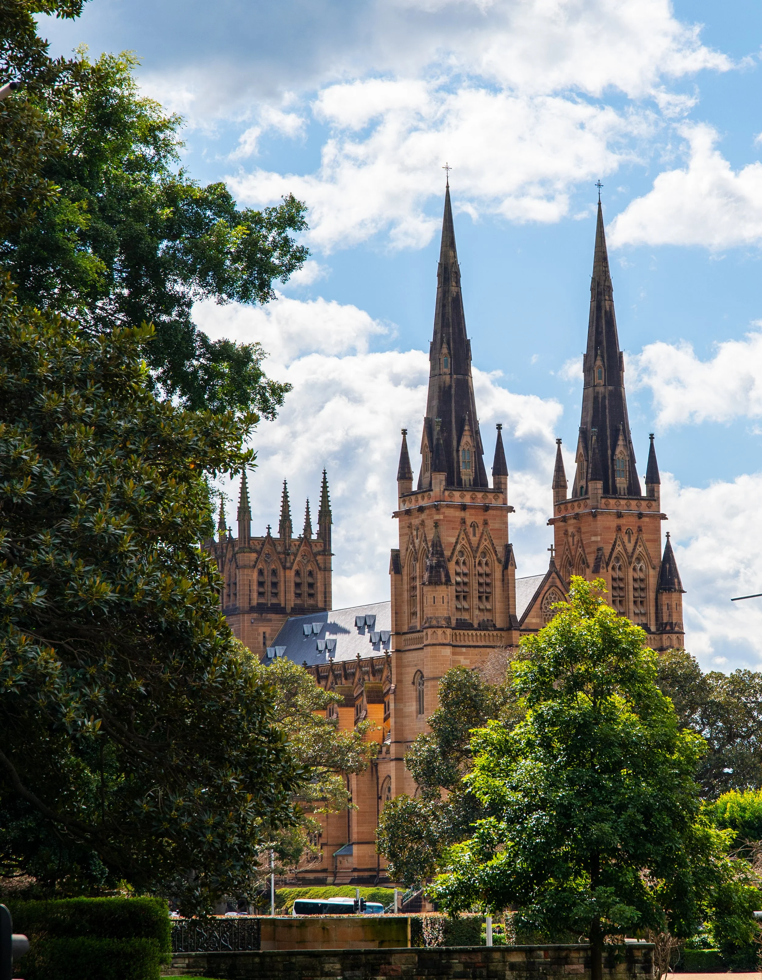 View of a large Gothic-style church with two tall spires, surrounded by trees and a partly cloudy sky.