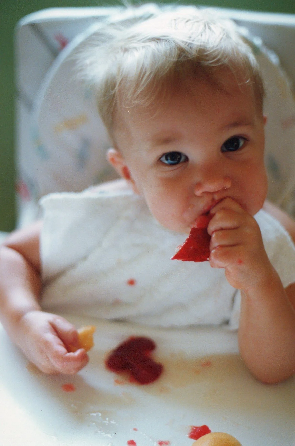 A young child with light hair in a high chair, eating strawberry slices with some fruit spread on the tray.