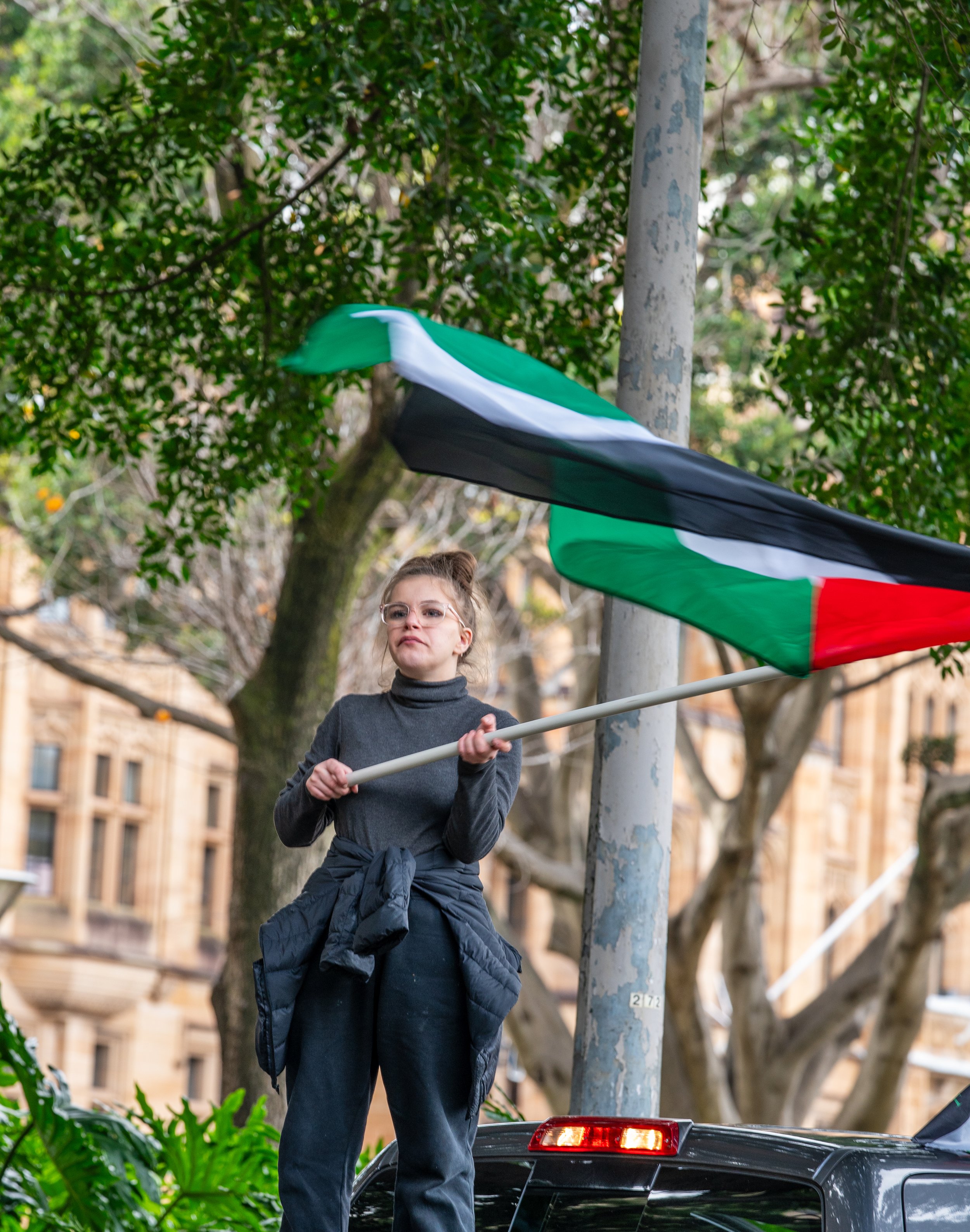 A woman holding a Palestinian flag standing on top of a car, in front of trees and buildings.