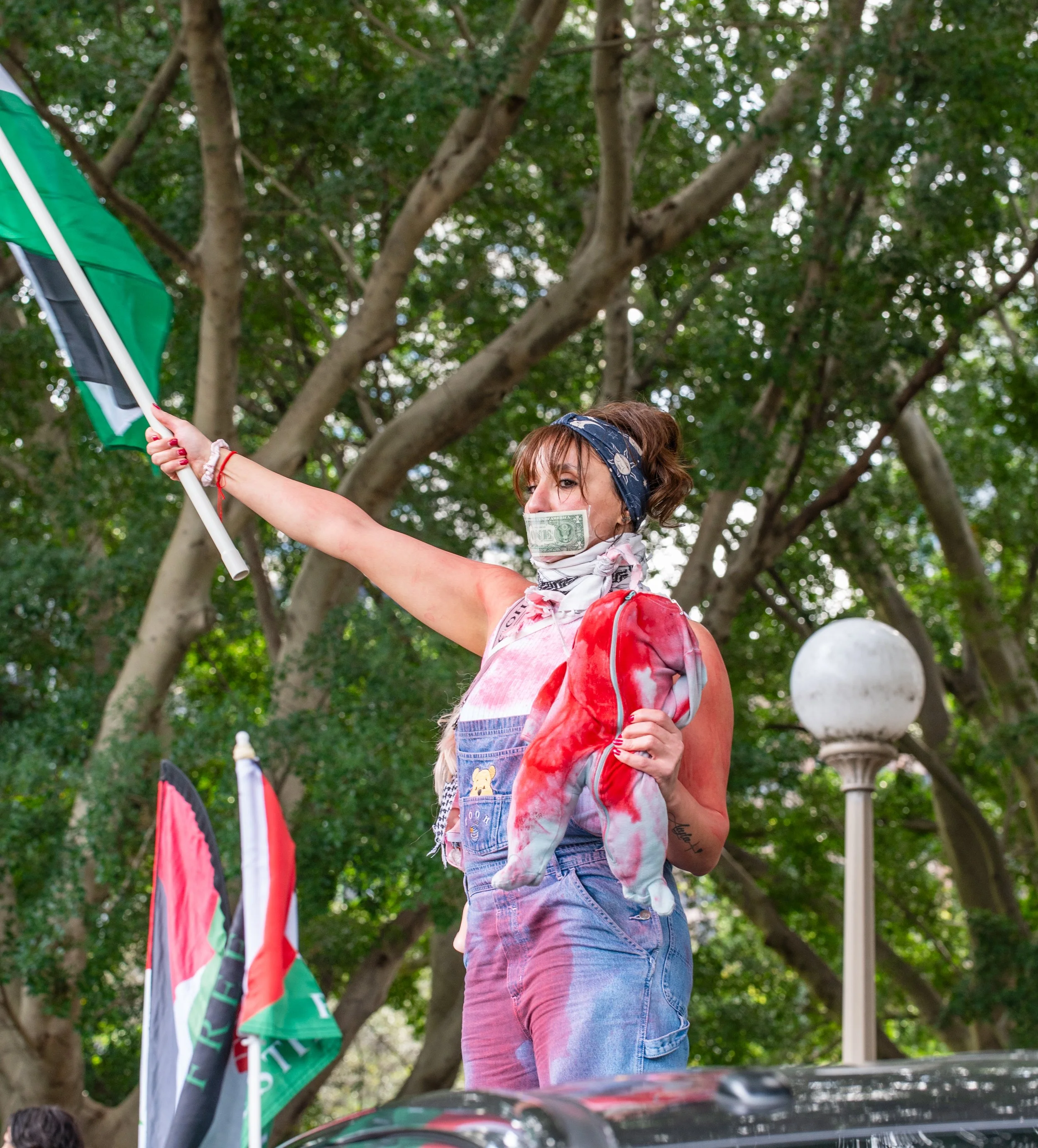 A woman with a dollar bill on her face, wearing a bandana and overalls, holding a flag and a plush toy, participating in a protest or demonstration outdoors among trees.