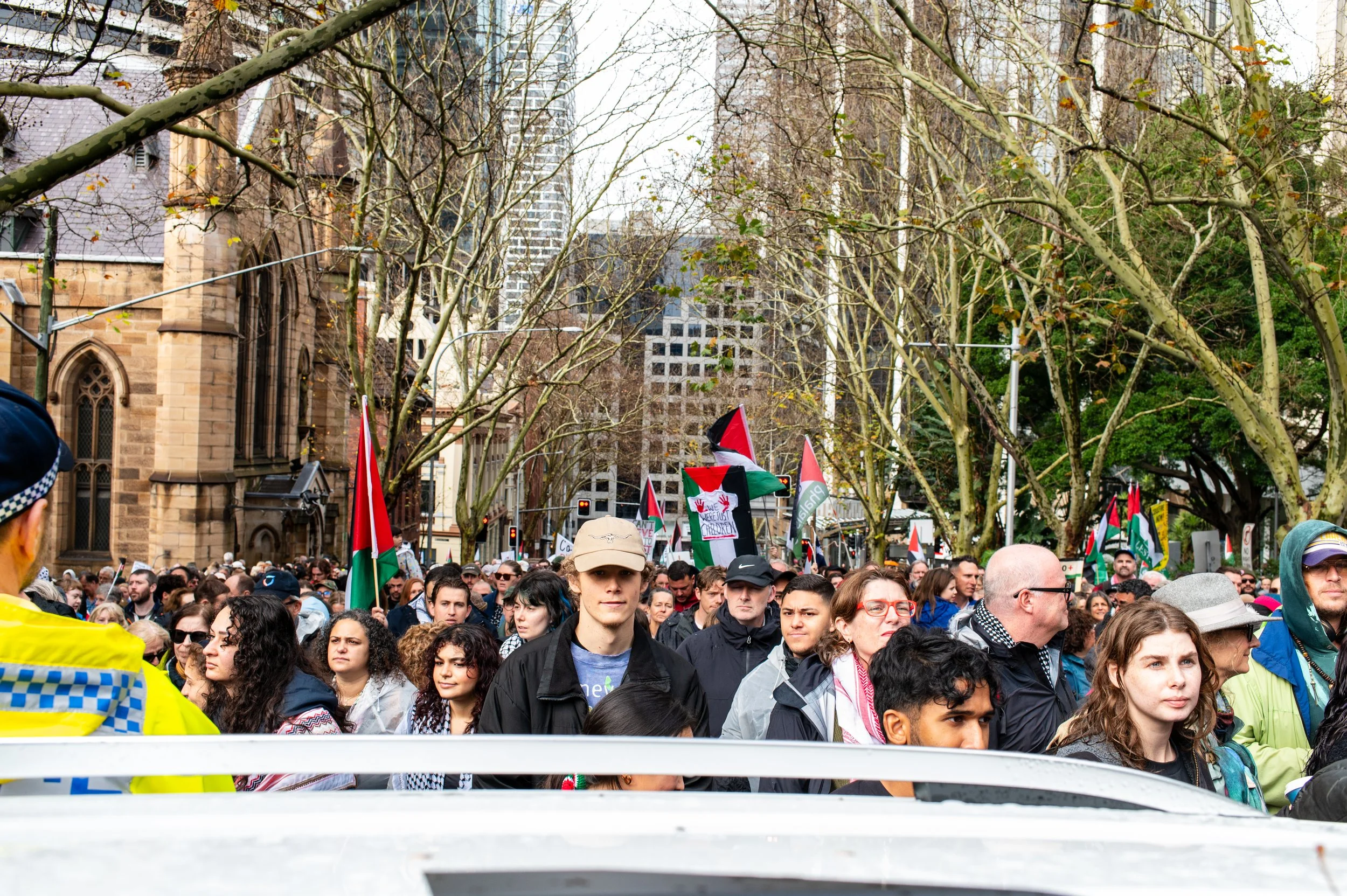 Crowd of people participating in a protest or rally under leafless trees in an urban area with tall buildings and a historic church in the background, some holding Palestinian flags.