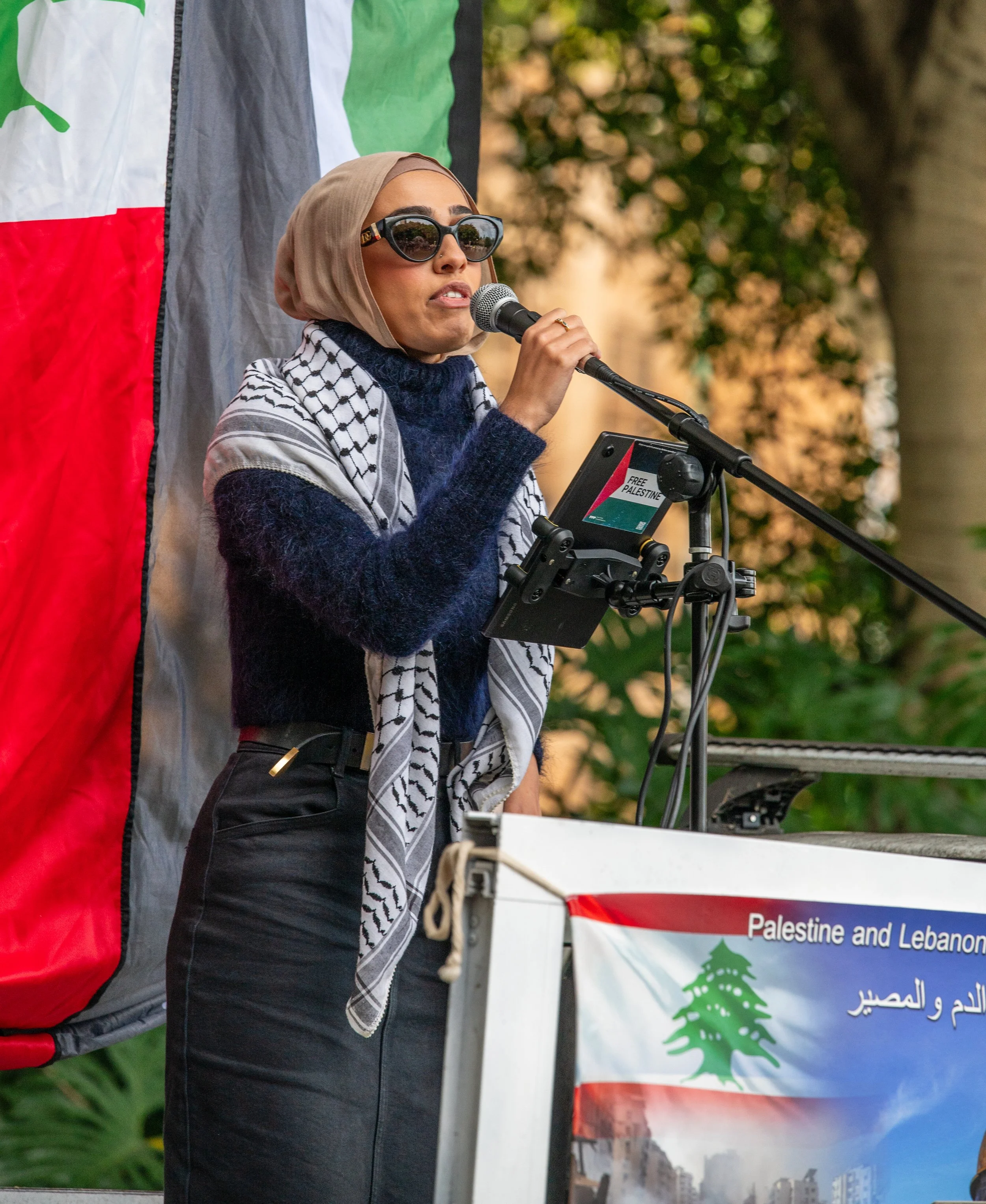 A woman wearing a beige hijab and black sunglasses is speaking into a microphone at a rally or protest. She stands near a banner with text about Palestine and Lebanon, and a flag featuring a green tree on a red, white, and blue background.