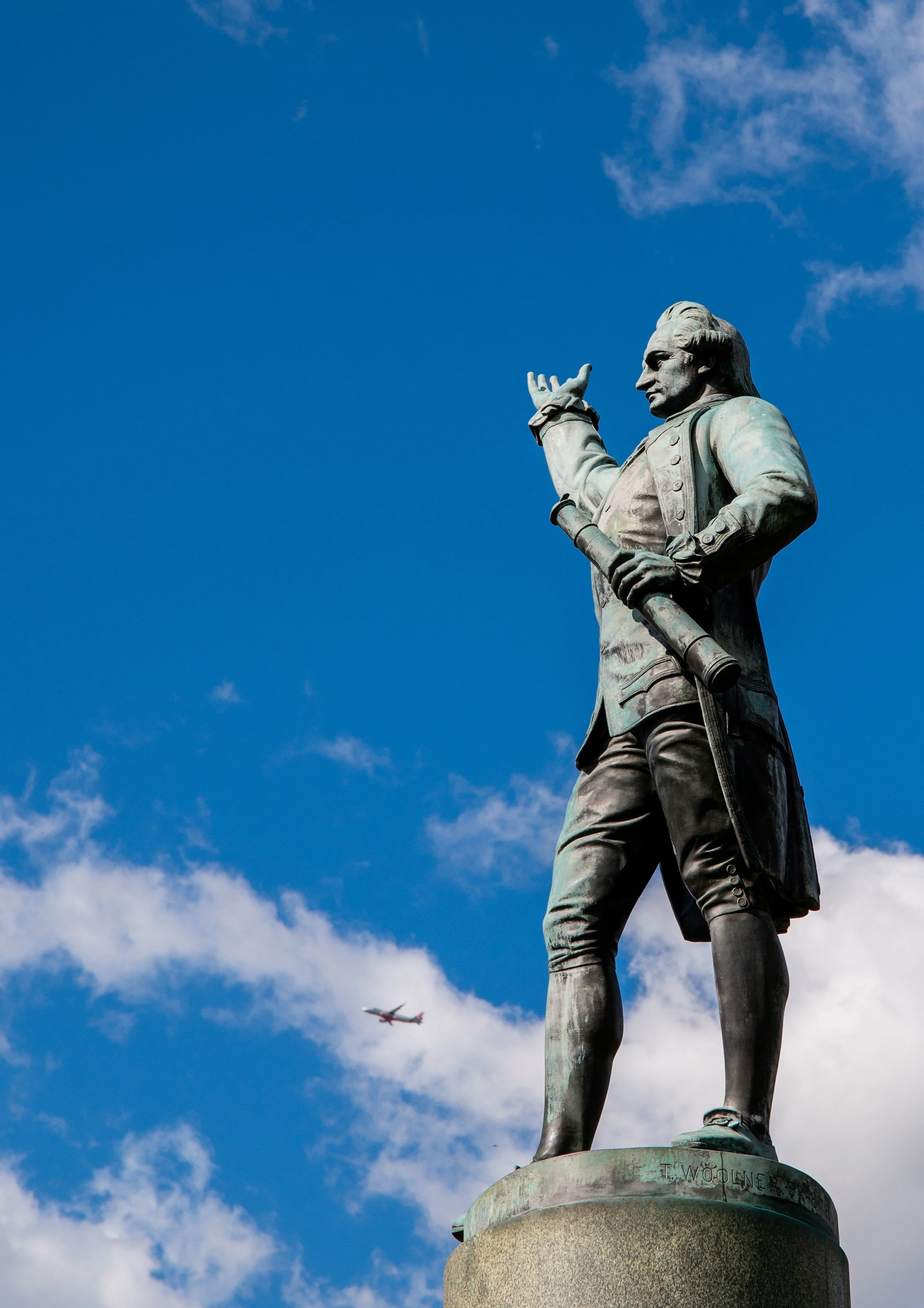Statue of a historical figure holding a telescope, standing on a pedestal against a blue sky with scattered clouds and an airplane flying in the background.