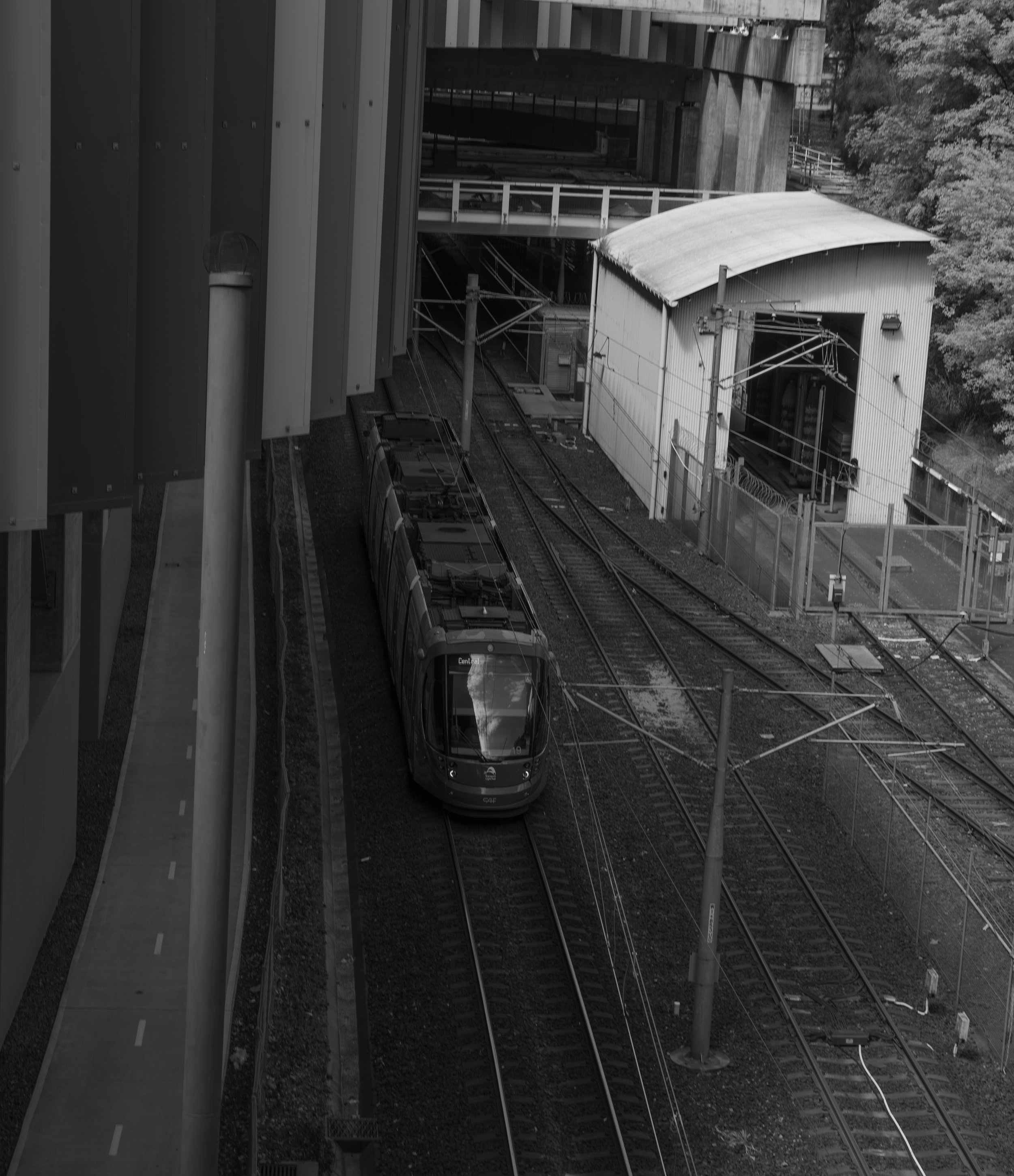 Black and white photo of a modern train on railway tracks next to an industrial building and a fenced area, with overhead wires and trees in the background.