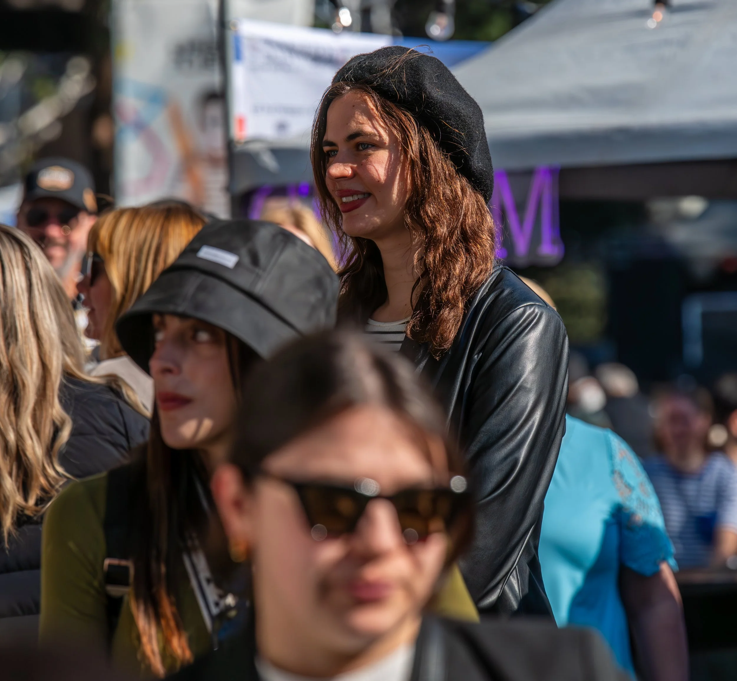 A diverse group of people gathered outdoors, with a woman with long brown hair, a black beret, and leather jacket standing out in focus, smiling among others.