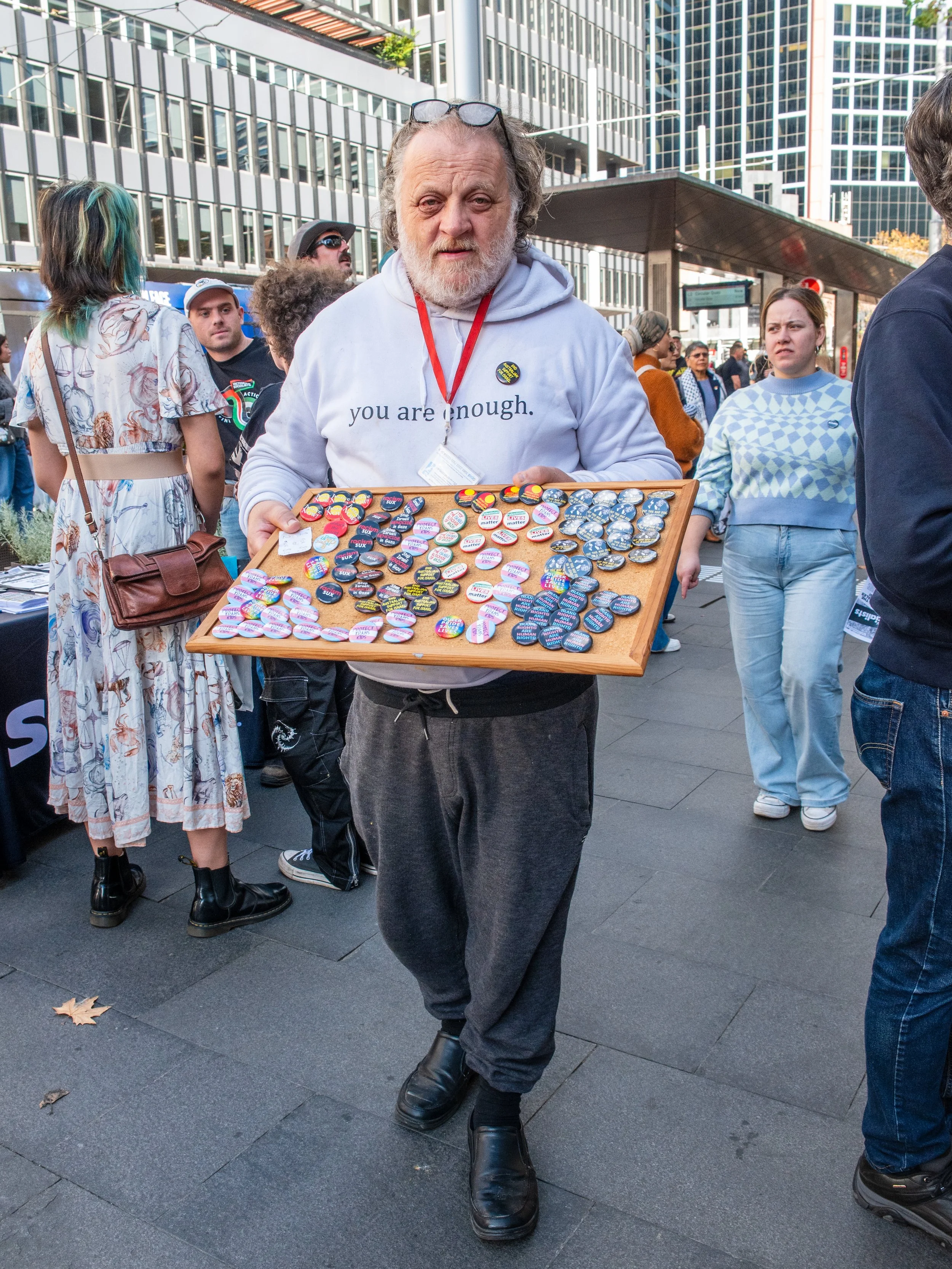 A man holding a display case filled with political buttons at an outdoor event in an urban area.