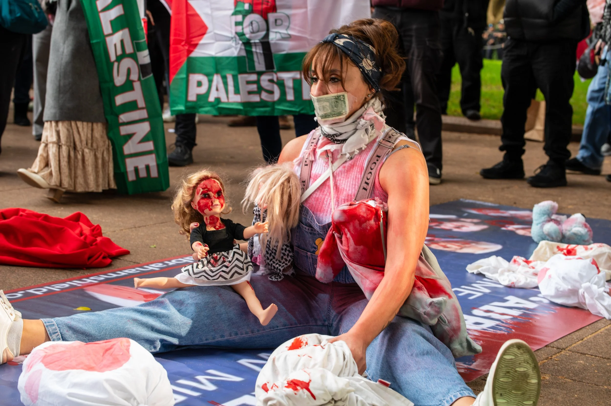 A woman sitting on the ground with a torn blue denim dress, bloodstained cloth, and a dollar bill over her face. She is surrounded by protest signs, dolls, and objects with blood stains, at a political demonstration with a Palestinian flag and other protestors in the background.