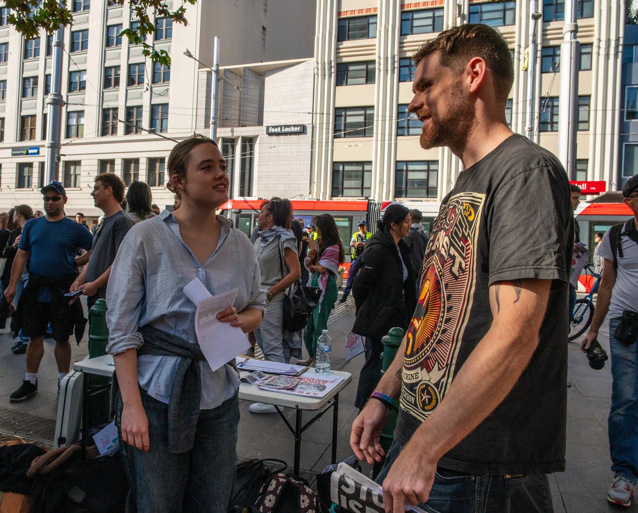 A young woman and a young man are having a conversation on a busy city street, possibly at a protest or event, with several people and buildings in the background.