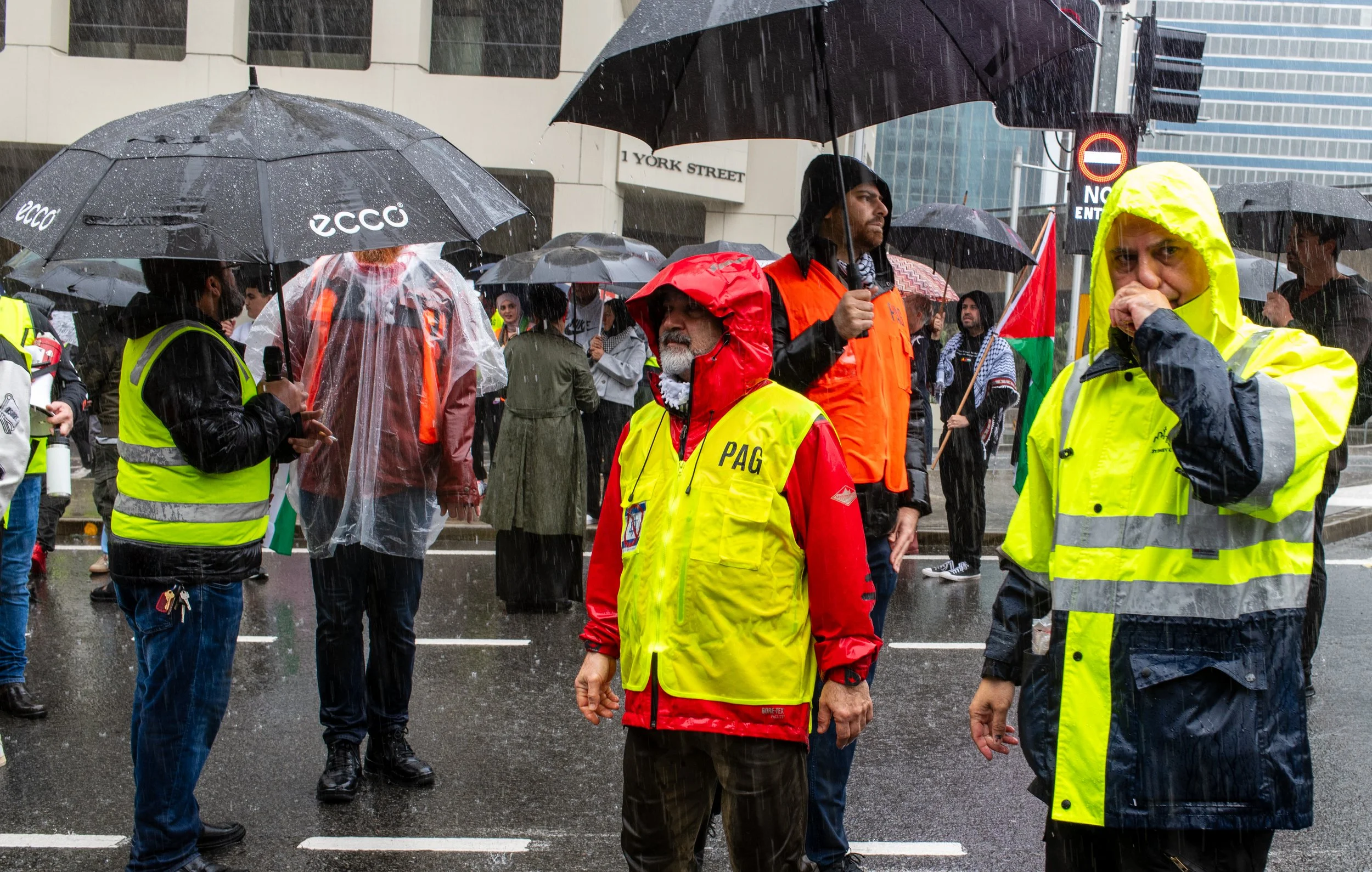 People standing in the rain during a protest or gathering in an urban area, holding umbrellas and wearing rain jackets, with some wearing yellow vests.