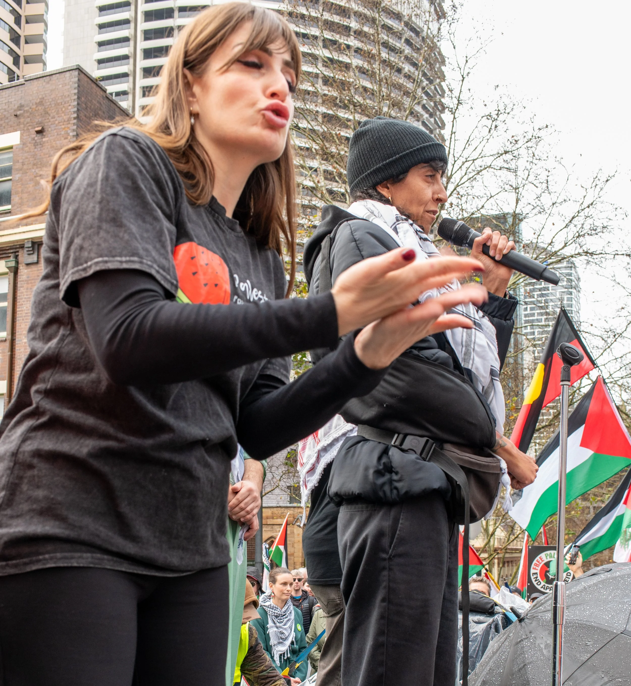 Two women speaking at a protest, with one holding a microphone and the other gesturing with her hands, in front of flags and a crowd of people with tall buildings in the background.