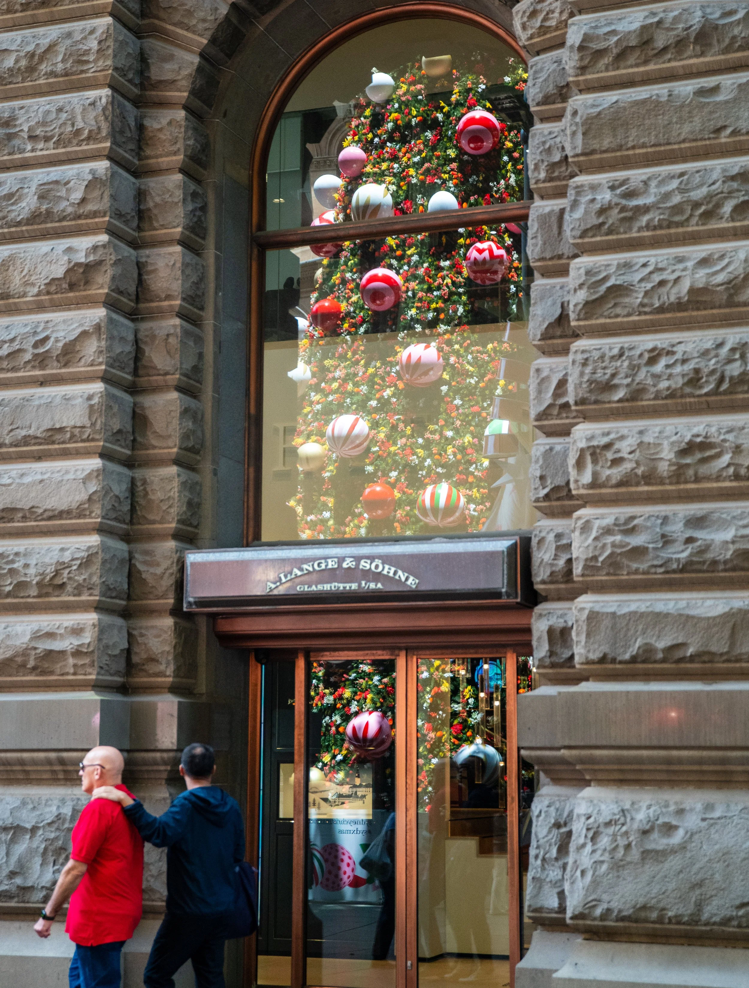 A large Christmas tree decorated with colorful ornaments is seen through a glass window of a building. The building has a stone exterior with a black sign that reads 'LANGE & SÖHNE.' Two men walk past in the foreground, one wearing a red shirt and the other in a dark jacket.