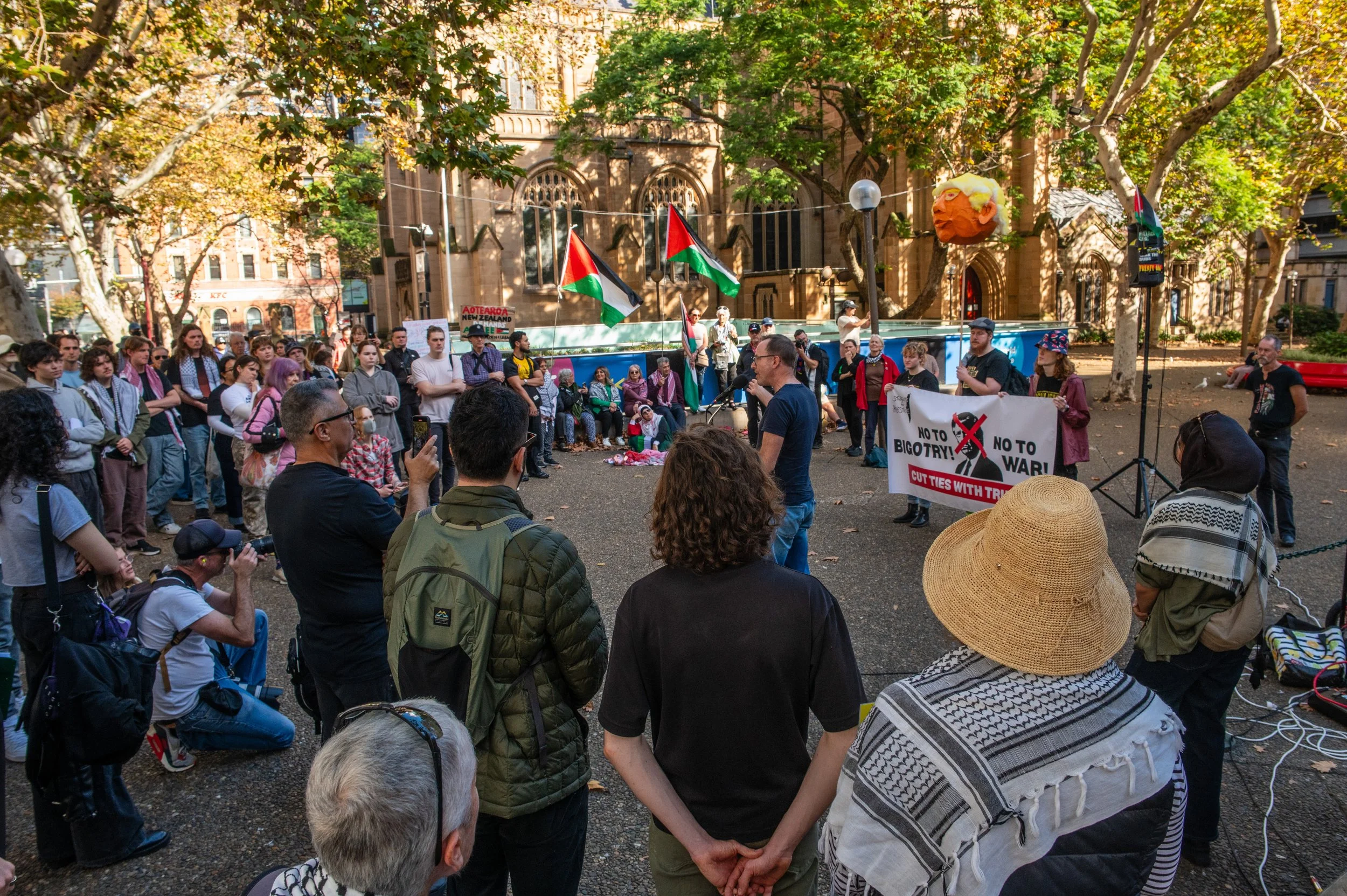 Protest rally in a park with a speaker at a microphone, holding a banner against bigotry and war, surrounded by a crowd of diverse people, some taking photos, with Palestinian flags and large trees providing shade.