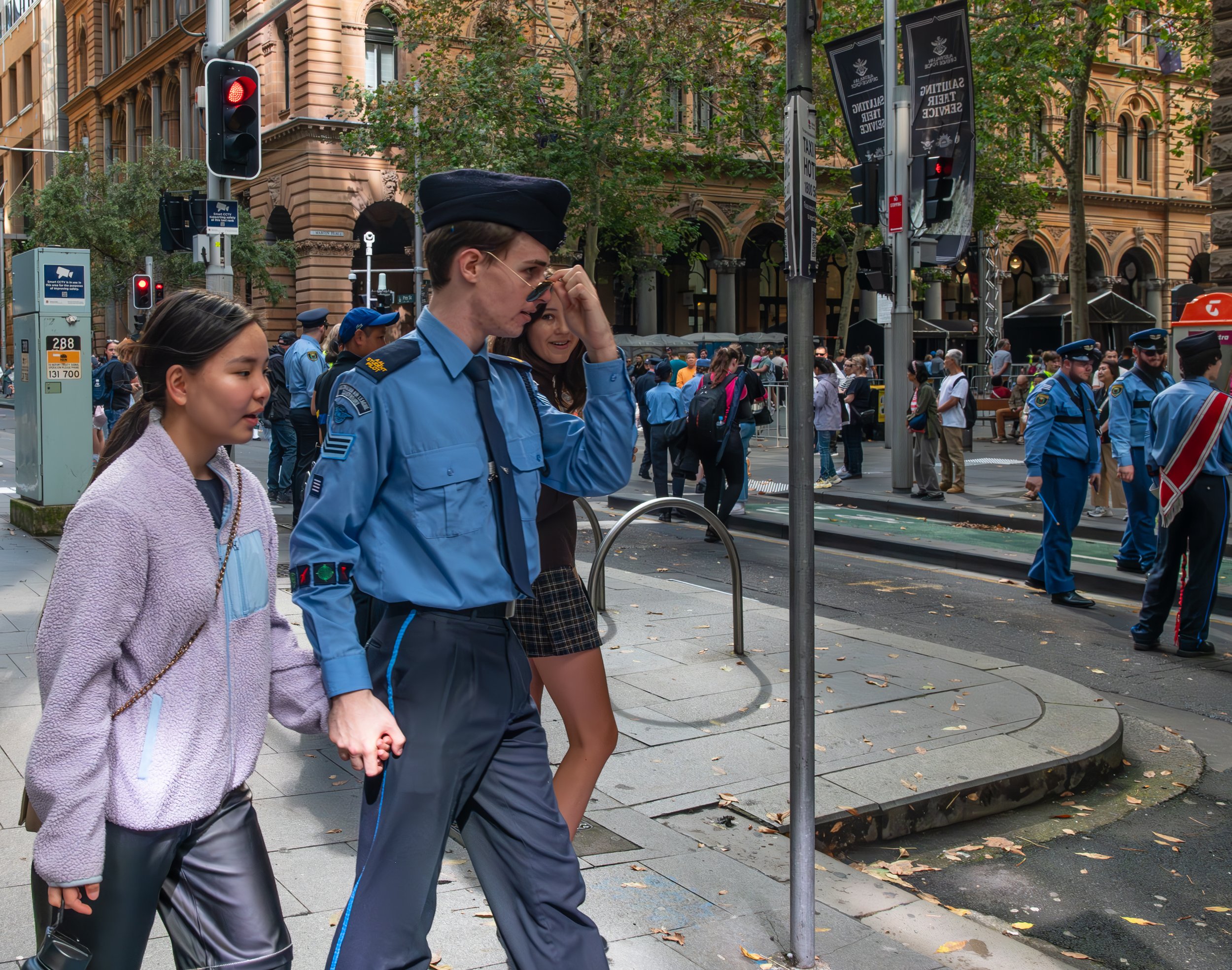 A police officer and two young women walk across a city street, with police and crowd in the background; streetlights and trees lining the street.